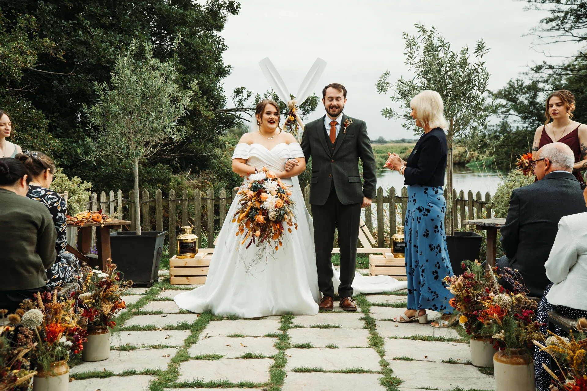 A happy bride holding an autumnal bouquet and a groom in a green suit stand together during an outdoor ceremony while guests applaud.
