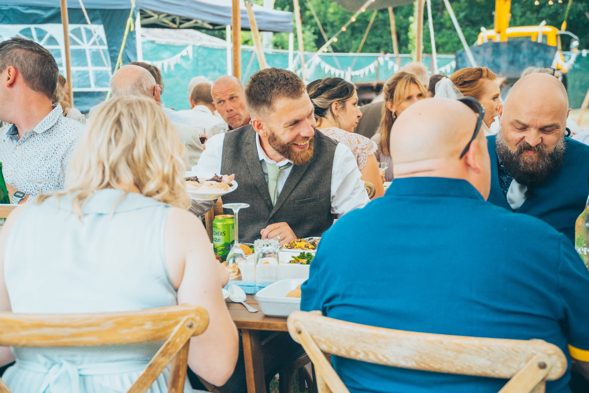 Wedding guests enjoy a lively wedding breakfast seated at long banquet tables inside a marquee.