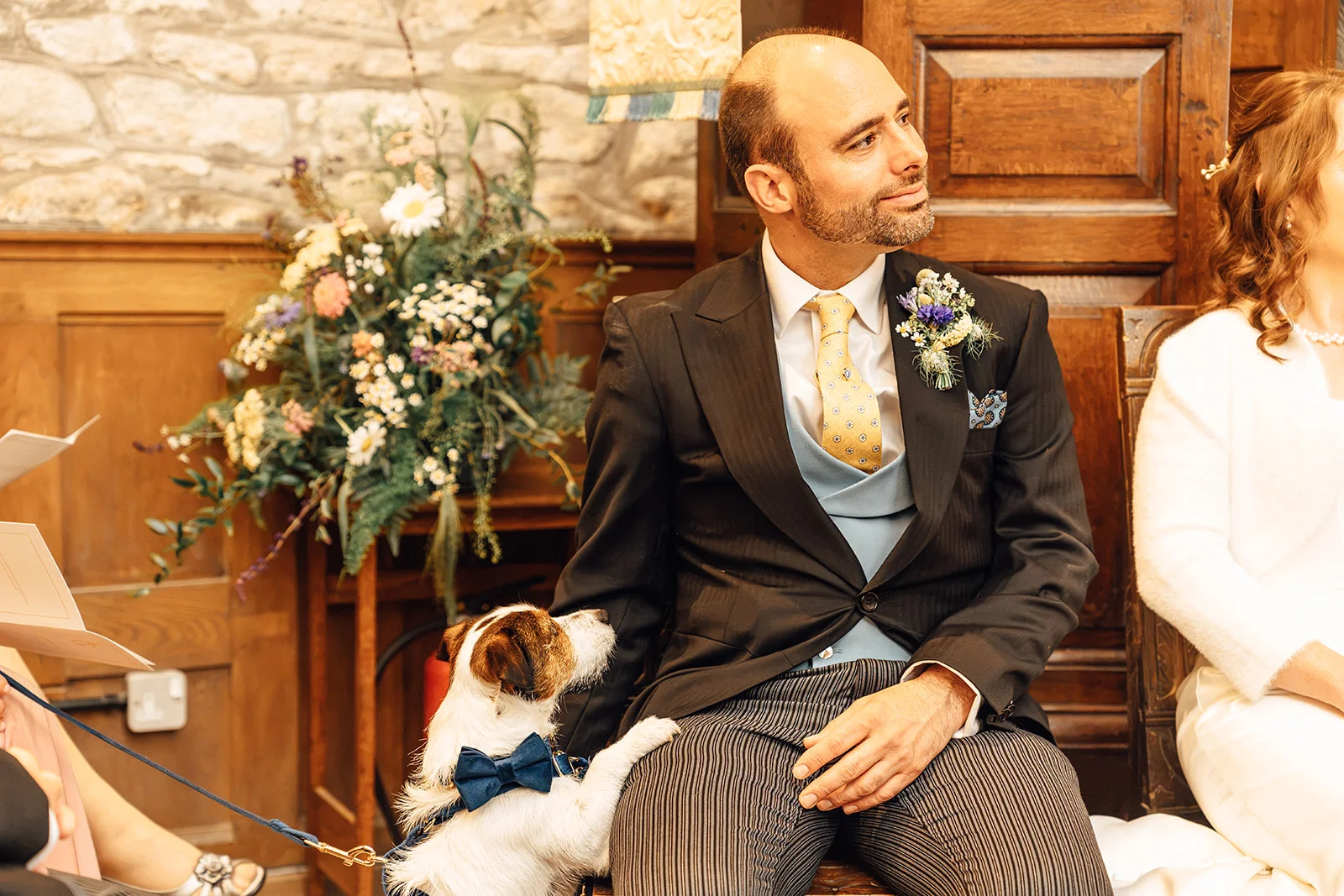 A groom in a traditional morning suit smiles during the ceremony while a Jack Russell terrier wearing a blue bow tie rests its paw on his knee.