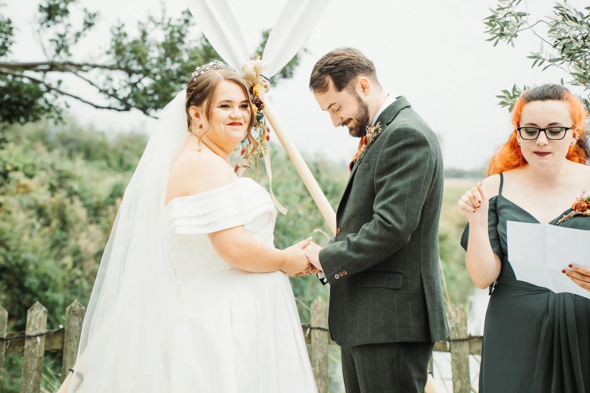A smiling bride and groom hold hands during an outdoor wedding ceremony, standing before a triangular wooden arch while a bridesmaid reads from a script nearby.