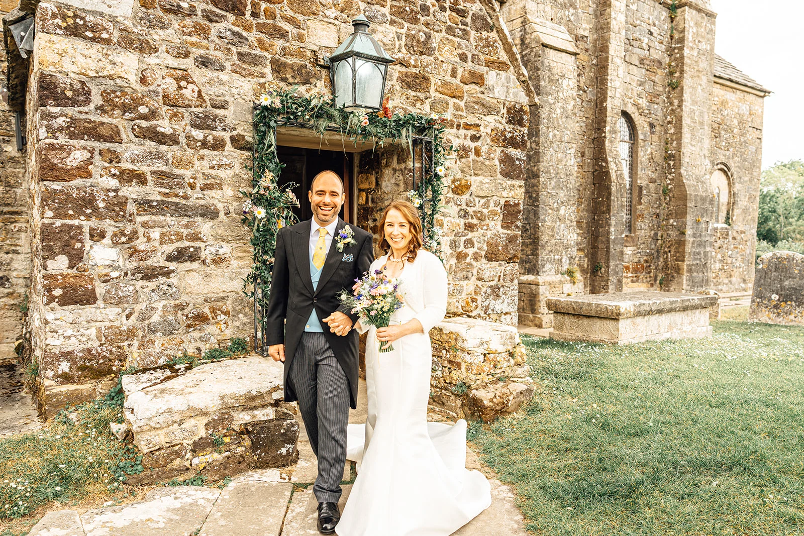 A newlywed couple smiling broadly as they walk hand-in-hand out of a historic stone church adorned with floral doorway decorations.