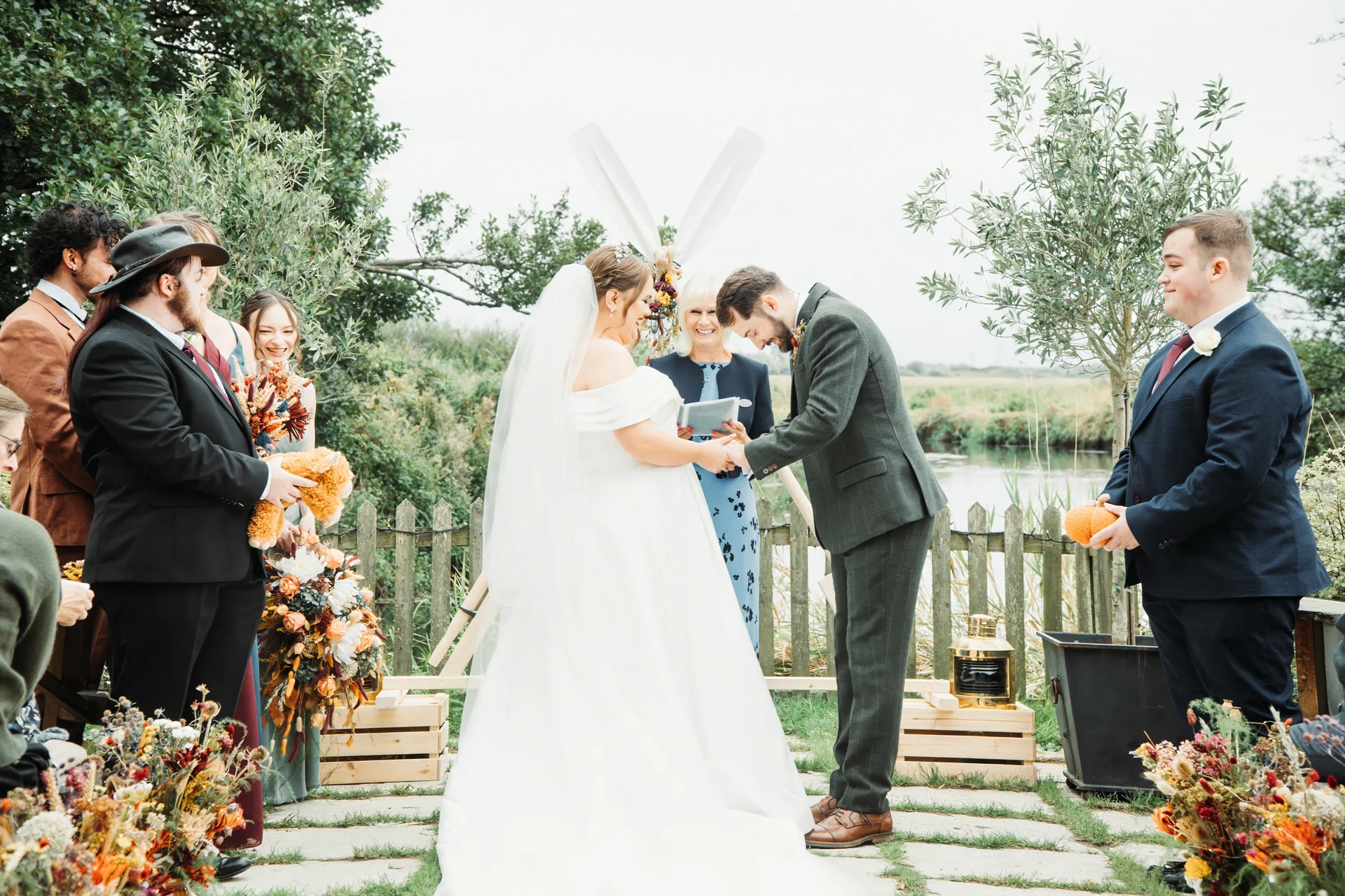 A bride in an off-the-shoulder gown and groom in a green suit exchange rings during an outdoor ceremony by a lake with family and bridal party watching.