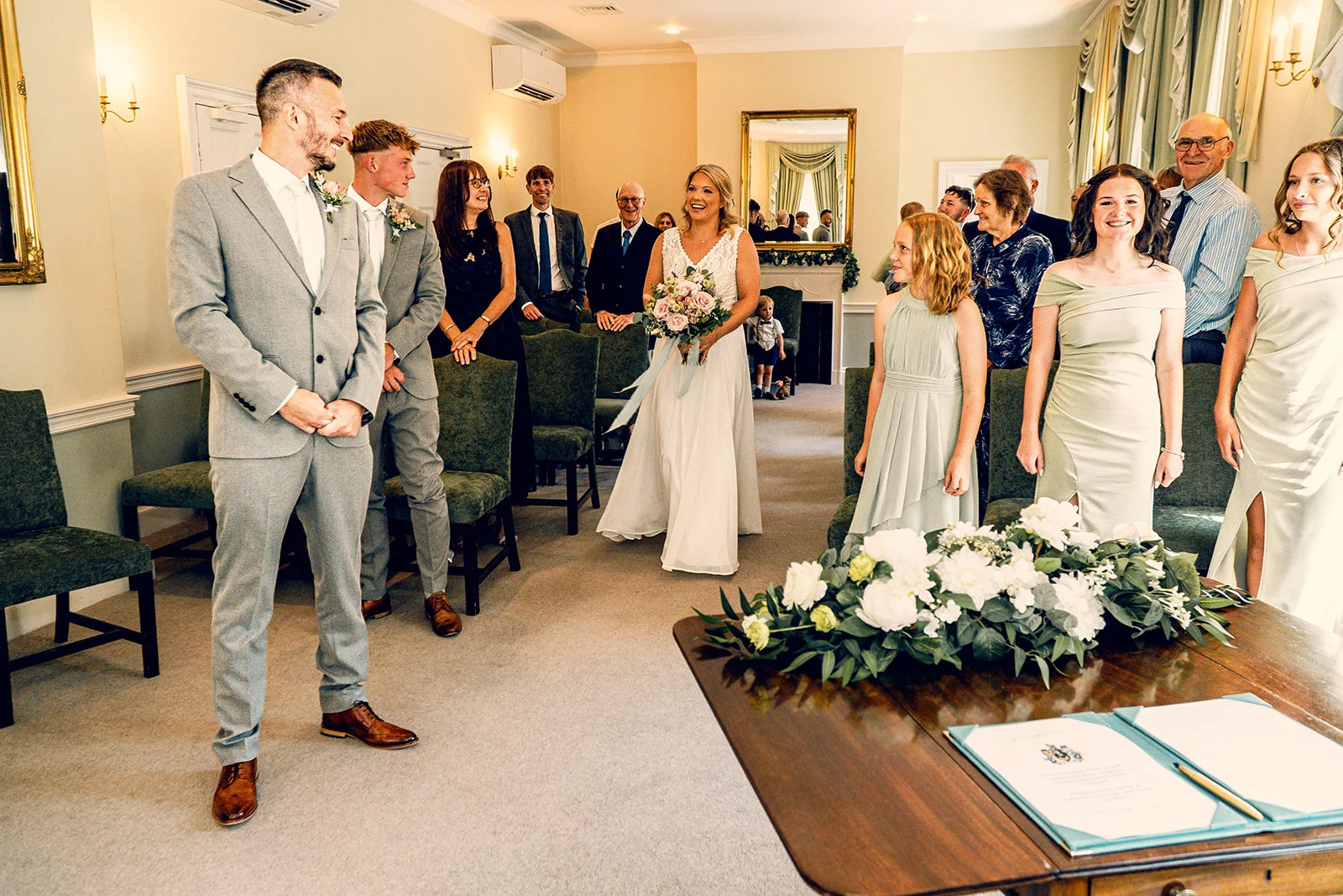 A smiling bride walks down the aisle holding a bouquet toward a groom waiting in a light grey suit during an indoor civil ceremony.