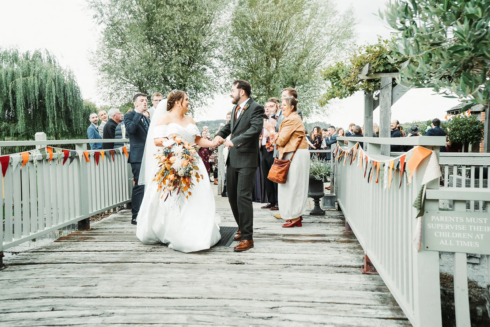 A newly married couple walk hand-in-hand across a wooden bridge decorated with bunting while cheering guests celebrate their union outdoors.