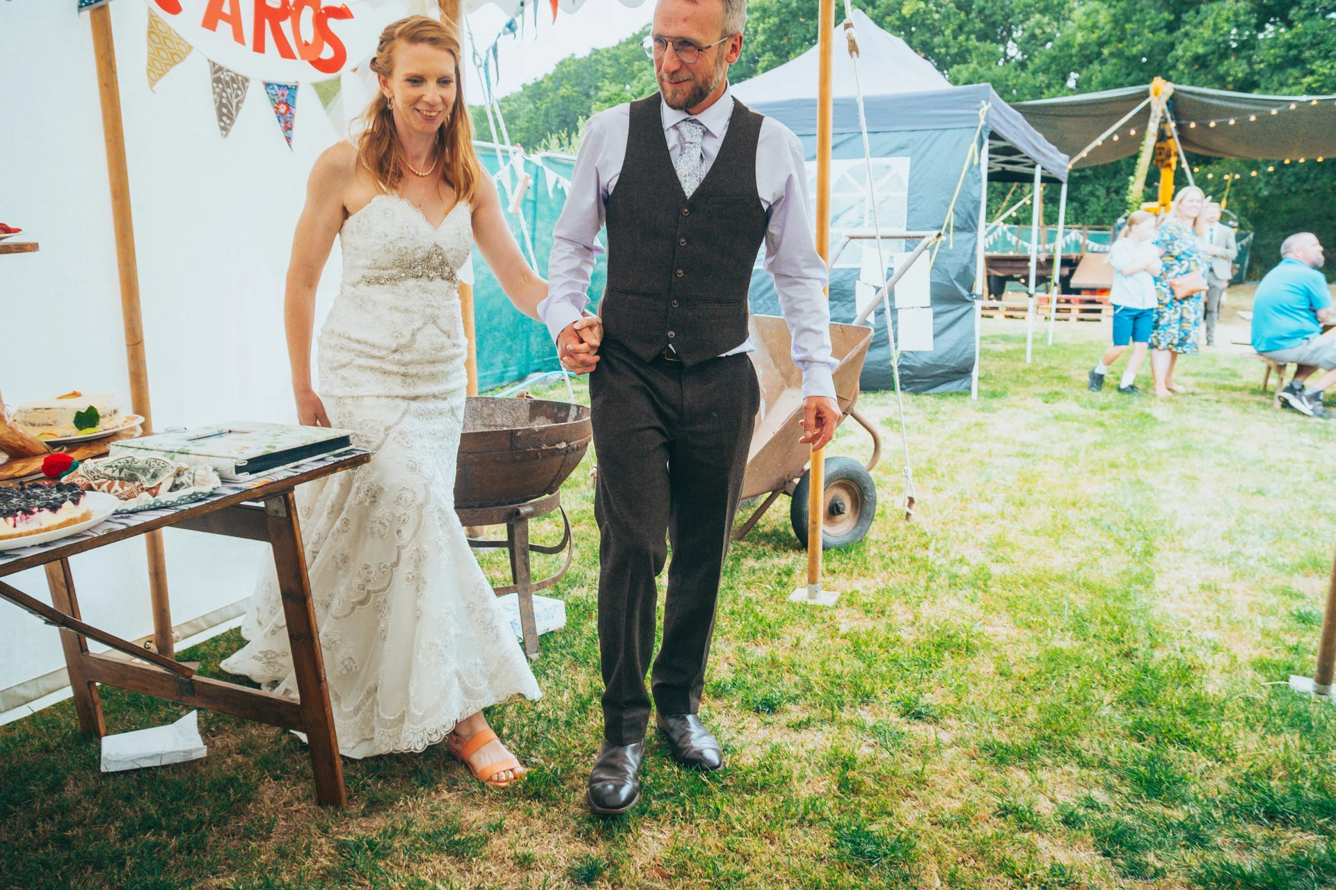 A newlywed couple walk hand in hand through an outdoor festival wedding reception with marquees and bunting in the background.