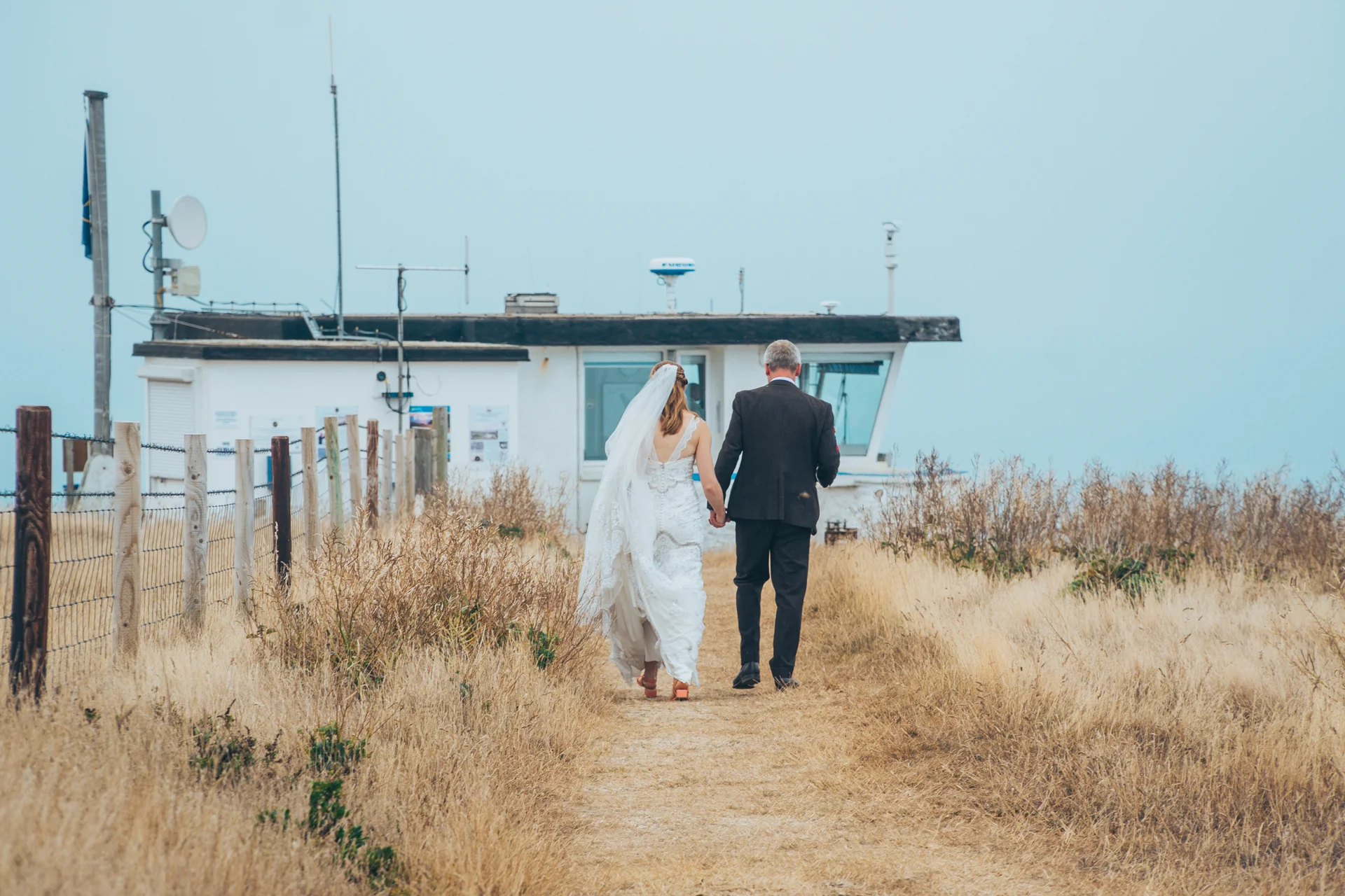 A newlywed couple walks hand in hand along a coastal path toward a lighthouse building under an overcast sky.