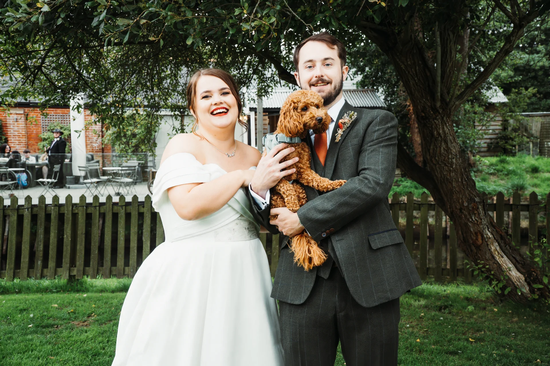 A smiling bride and groom pose outdoors in a lush garden setting while holding their fluffy brown pet dog between them.