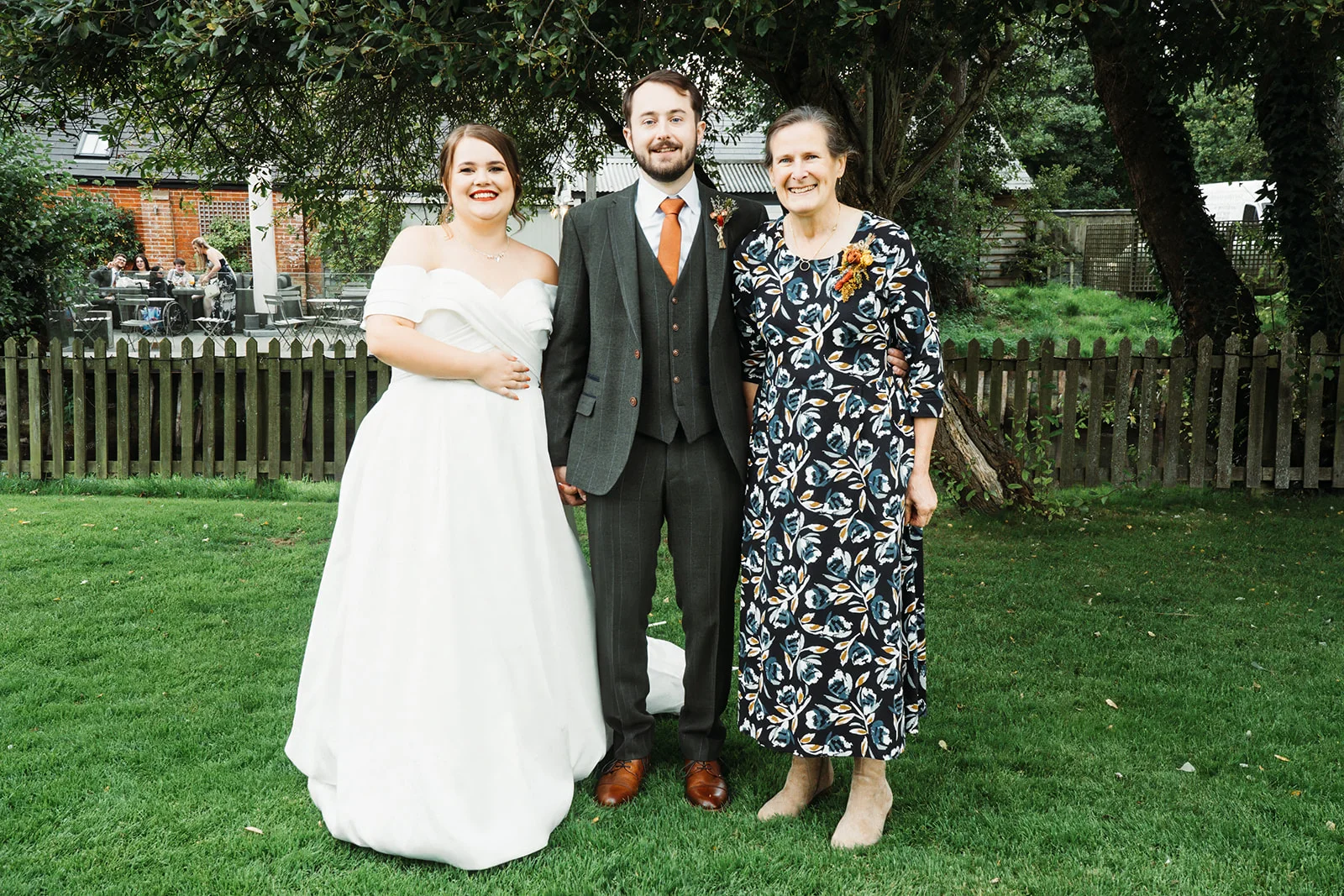 The bride in a white off-the-shoulder gown, the groom in a grey three-piece suit with an orange tie, and a guest in a floral dress pose together on a green lawn outdoors.