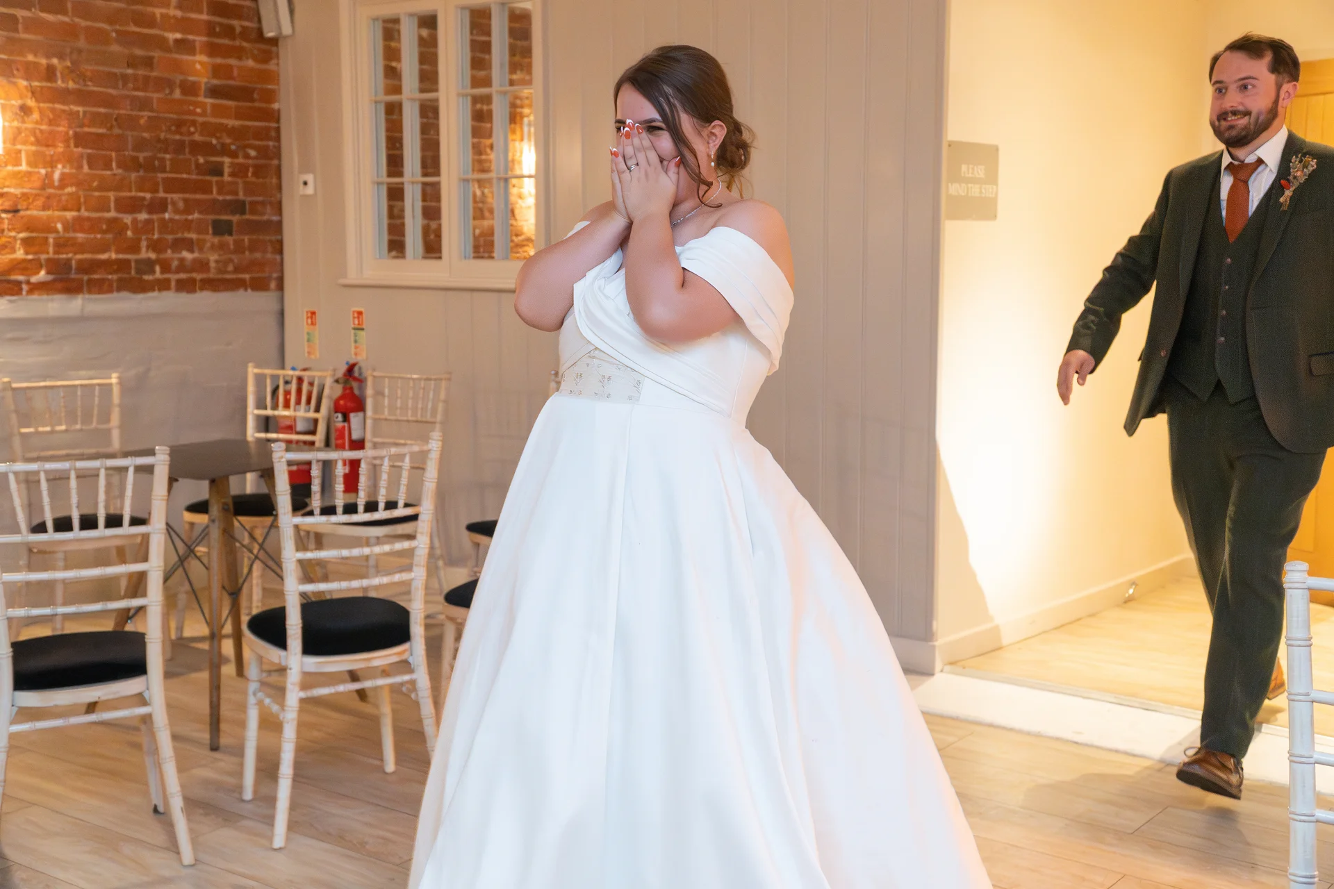 An emotional bride covers her face in tears of joy during a first look while the groom enters the room wearing a green tweed suit.