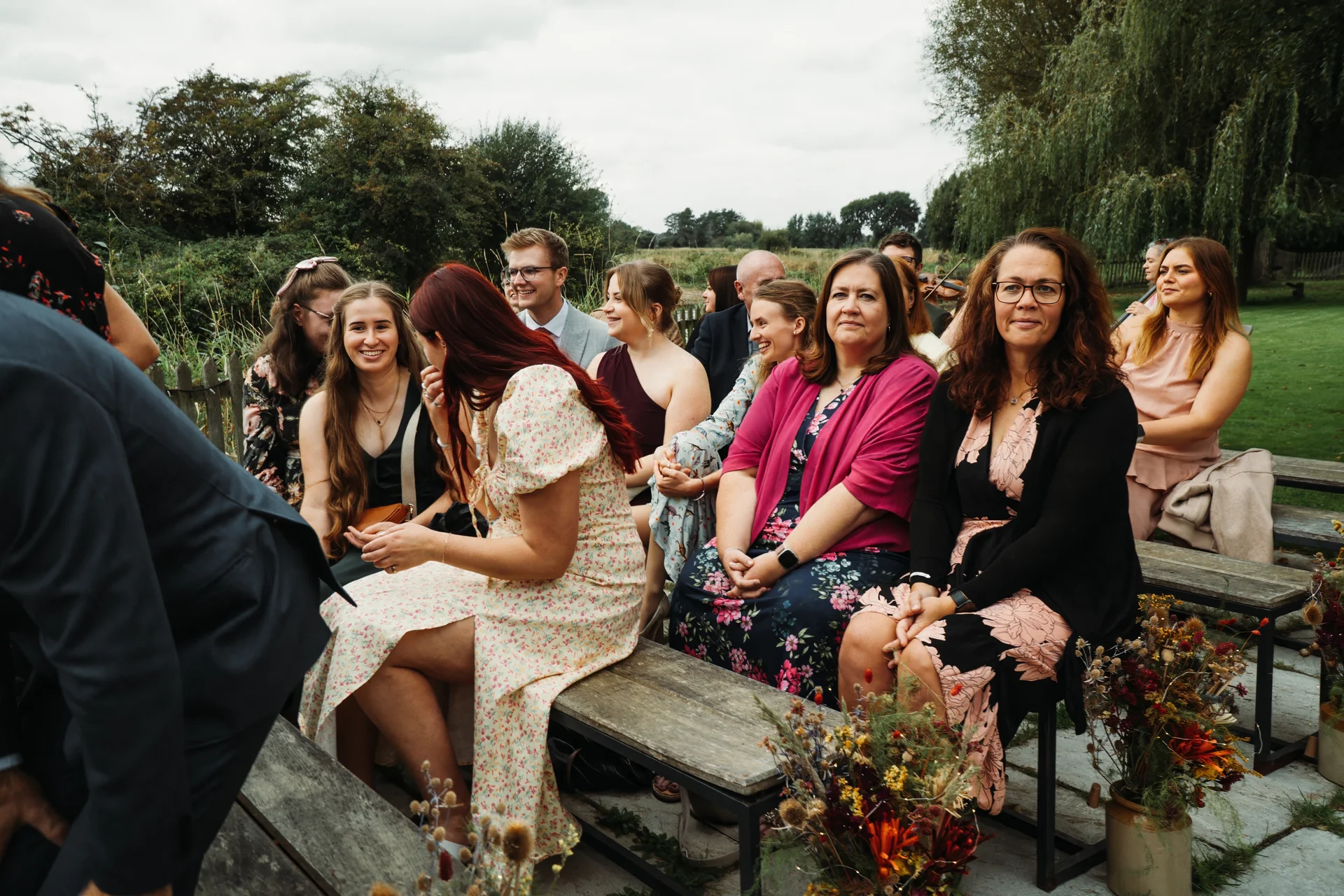 A candid group of well-dressed wedding guests seated on wooden benches outdoors, smiling and laughing during an outdoor ceremony.