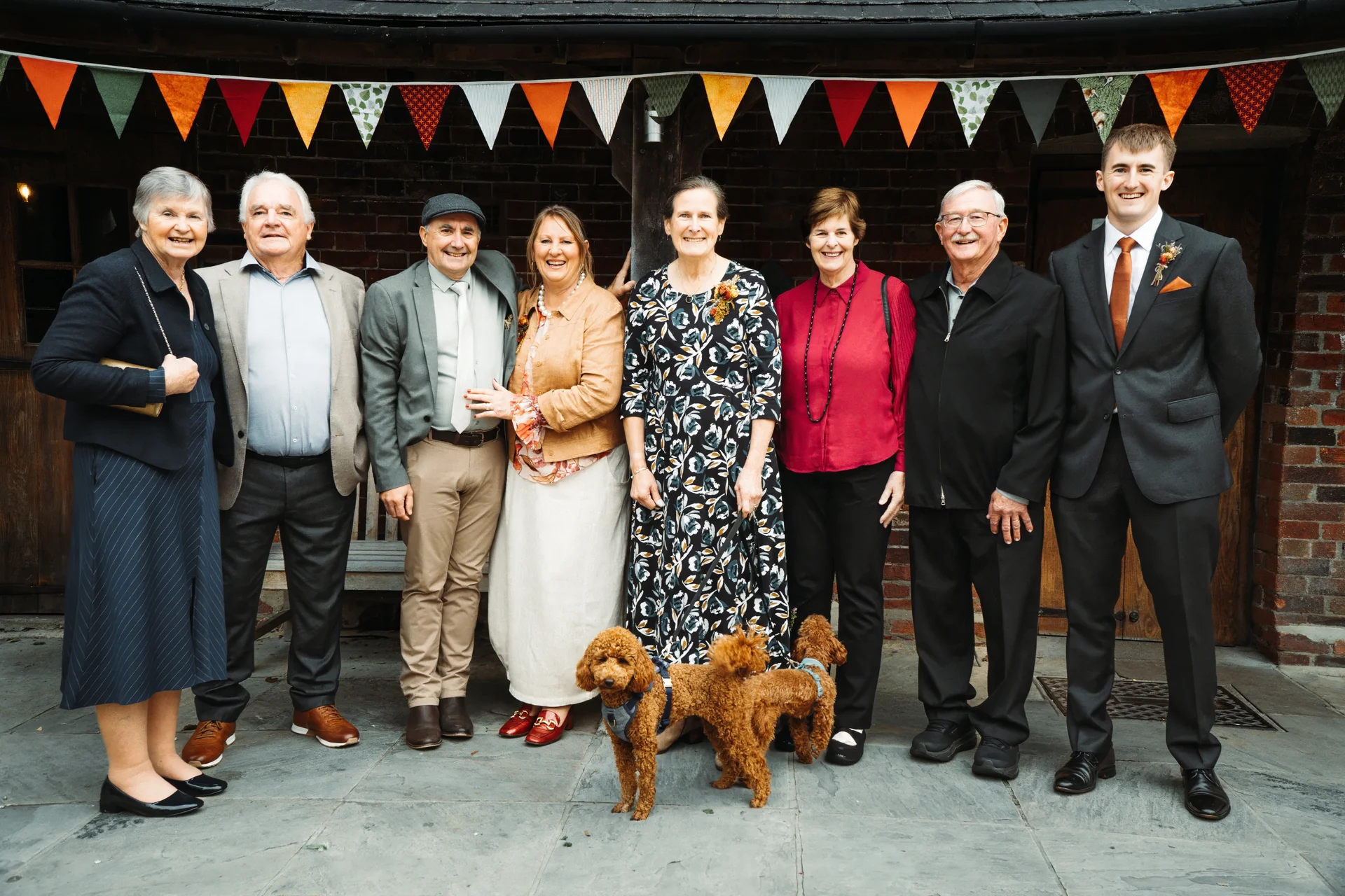 A group of family members and three small dogs pose together outdoors in front of a rustic brick wall decorated with colorful bunting.