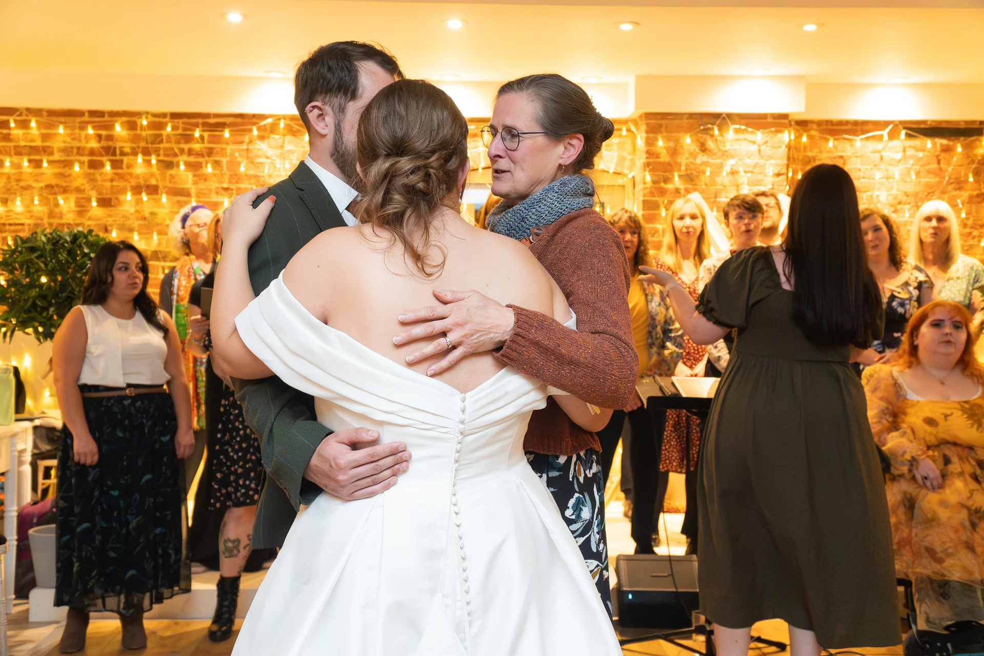 A bride in an off-the-shoulder satin gown and groom share a warm, emotional embrace with a guest in front of an exposed brick wall decorated with string lights.