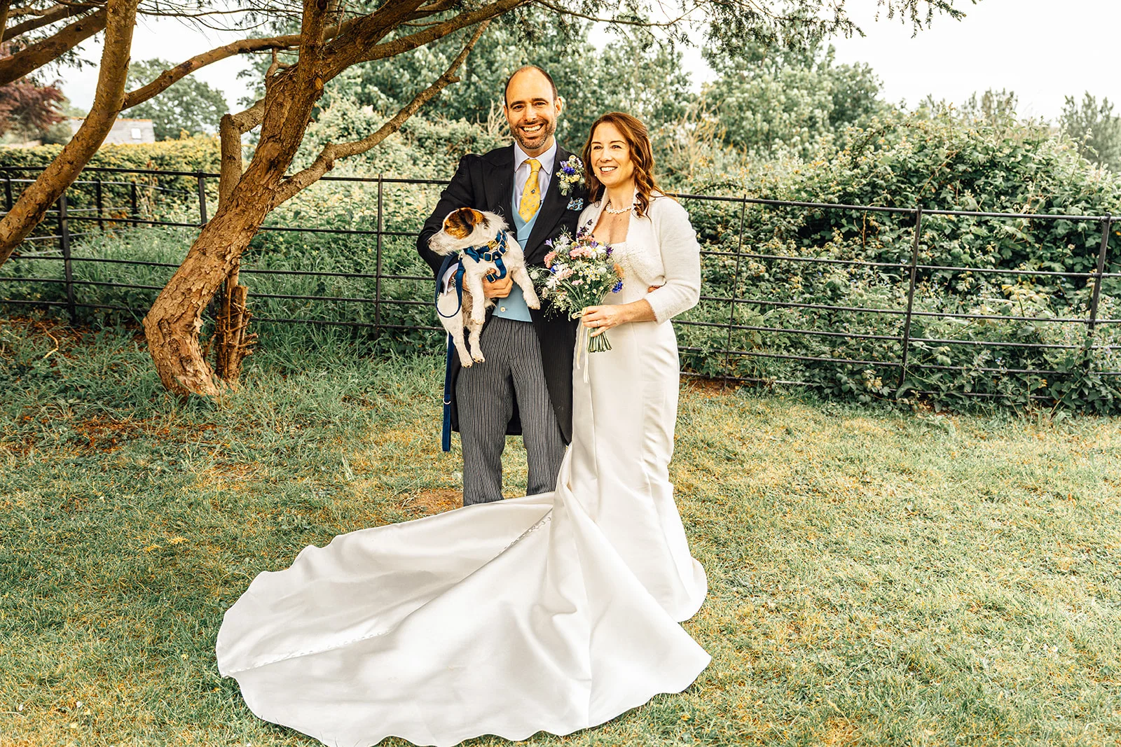 A smiling bride and groom pose outdoors in a lush garden setting, with the groom holding a small Jack Russell terrier while the bride holds a bouquet and showcases her long satin gown with a dramatic train.