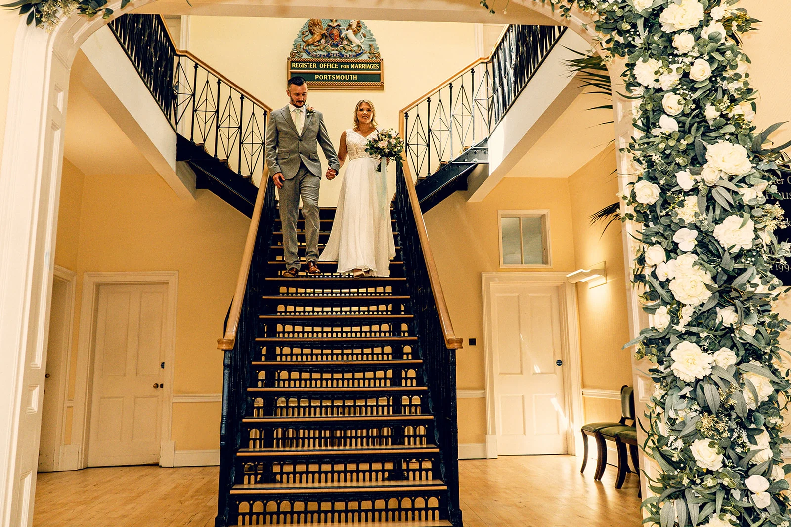 A newlywed couple walks hand in hand down a grand staircase at a Portsmouth register office, framed by a lush white floral arch.