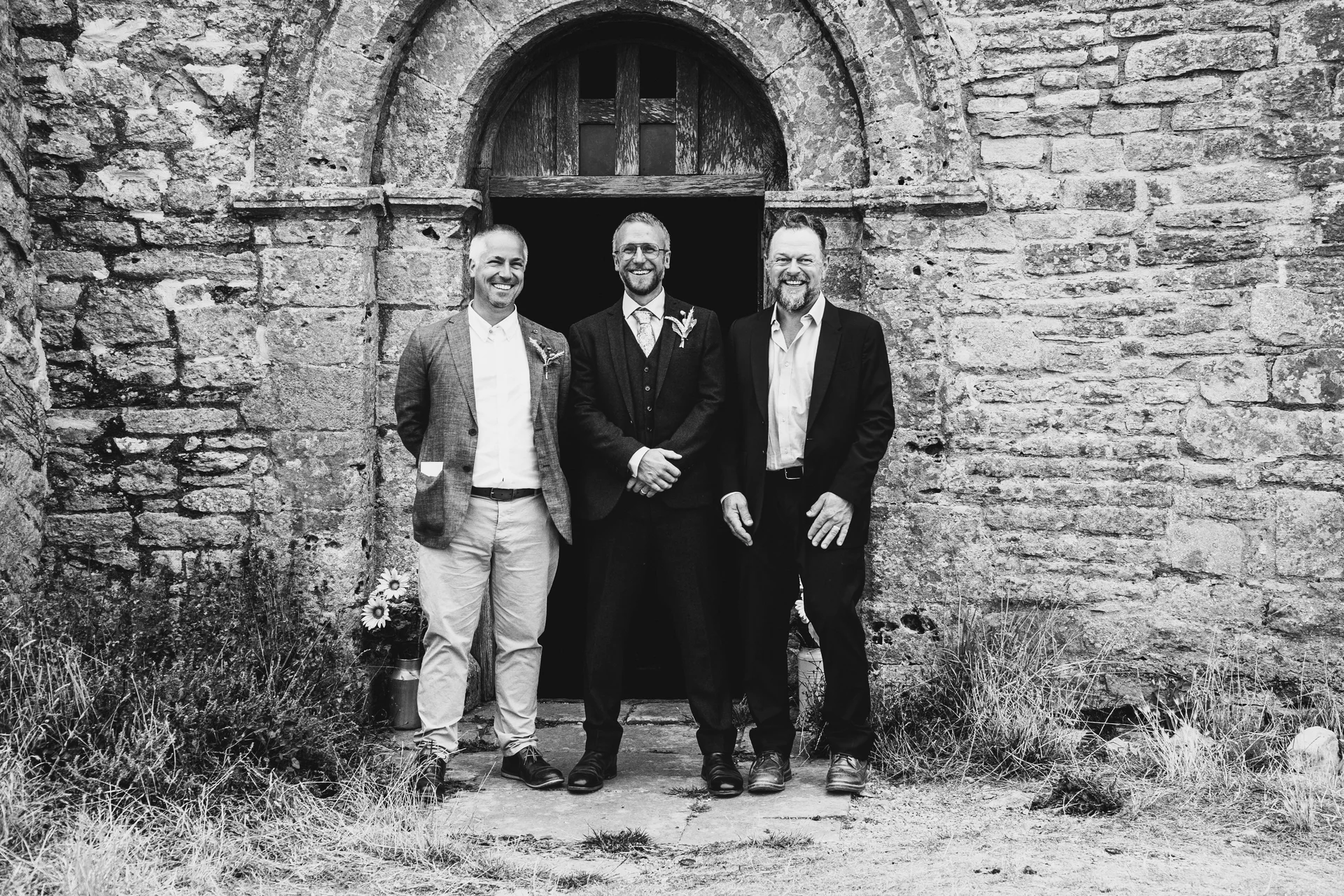 Three smiling men pose together outside an ancient stone building with a rustic wooden doorway.