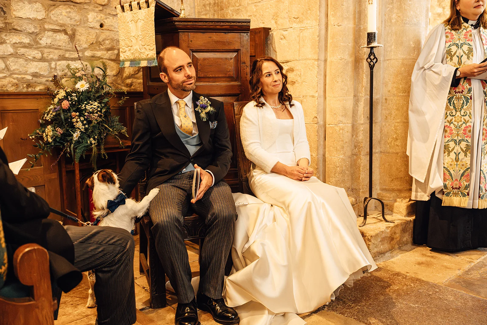 A bride in a satin gown and white stole sits beside her groom in a morning suit inside a historic stone church while a small dog with a blue bow tie places a paw on his knee.