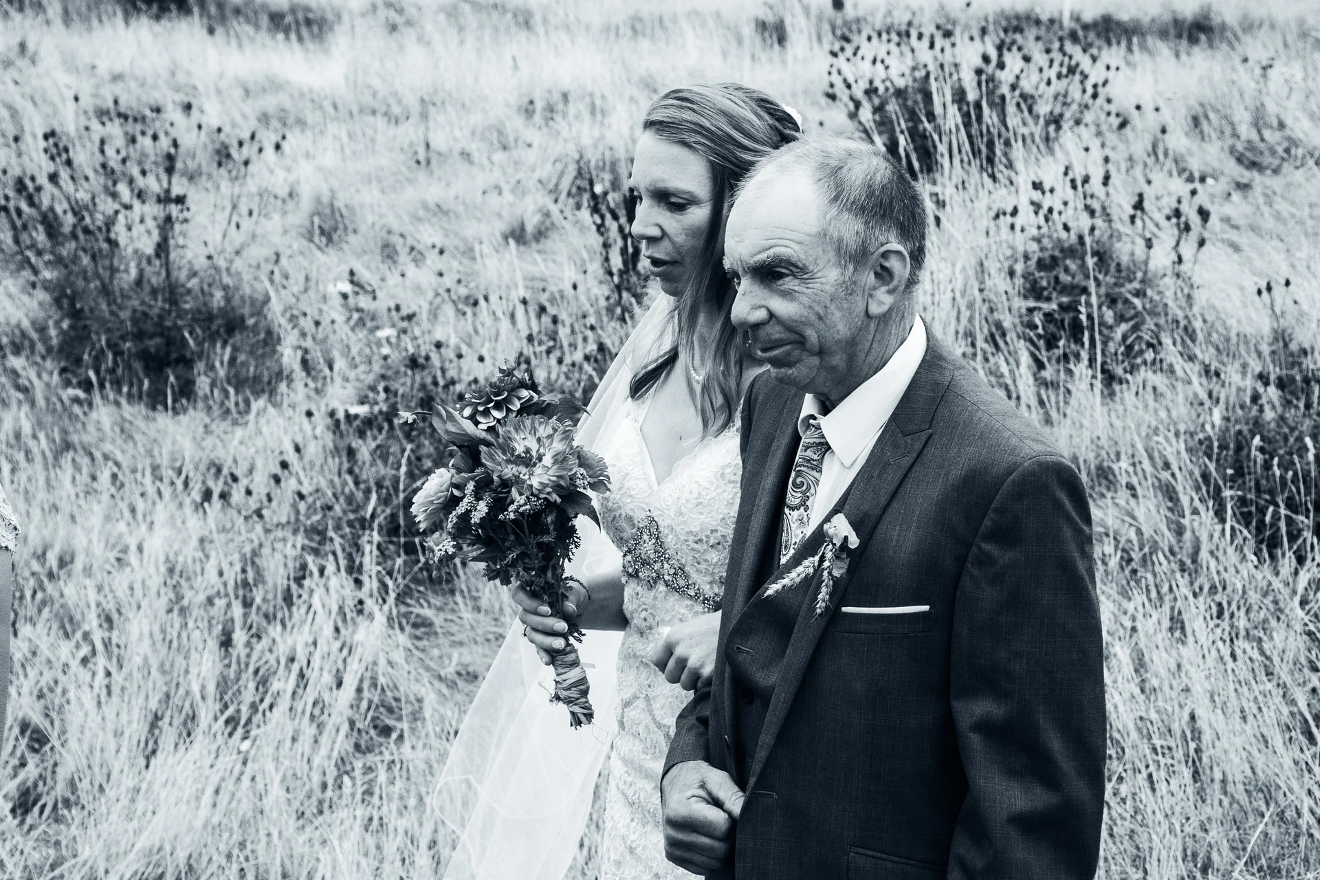 A bride holding a bouquet walks through a wildflower meadow with her father in a black and white documentary photograph.