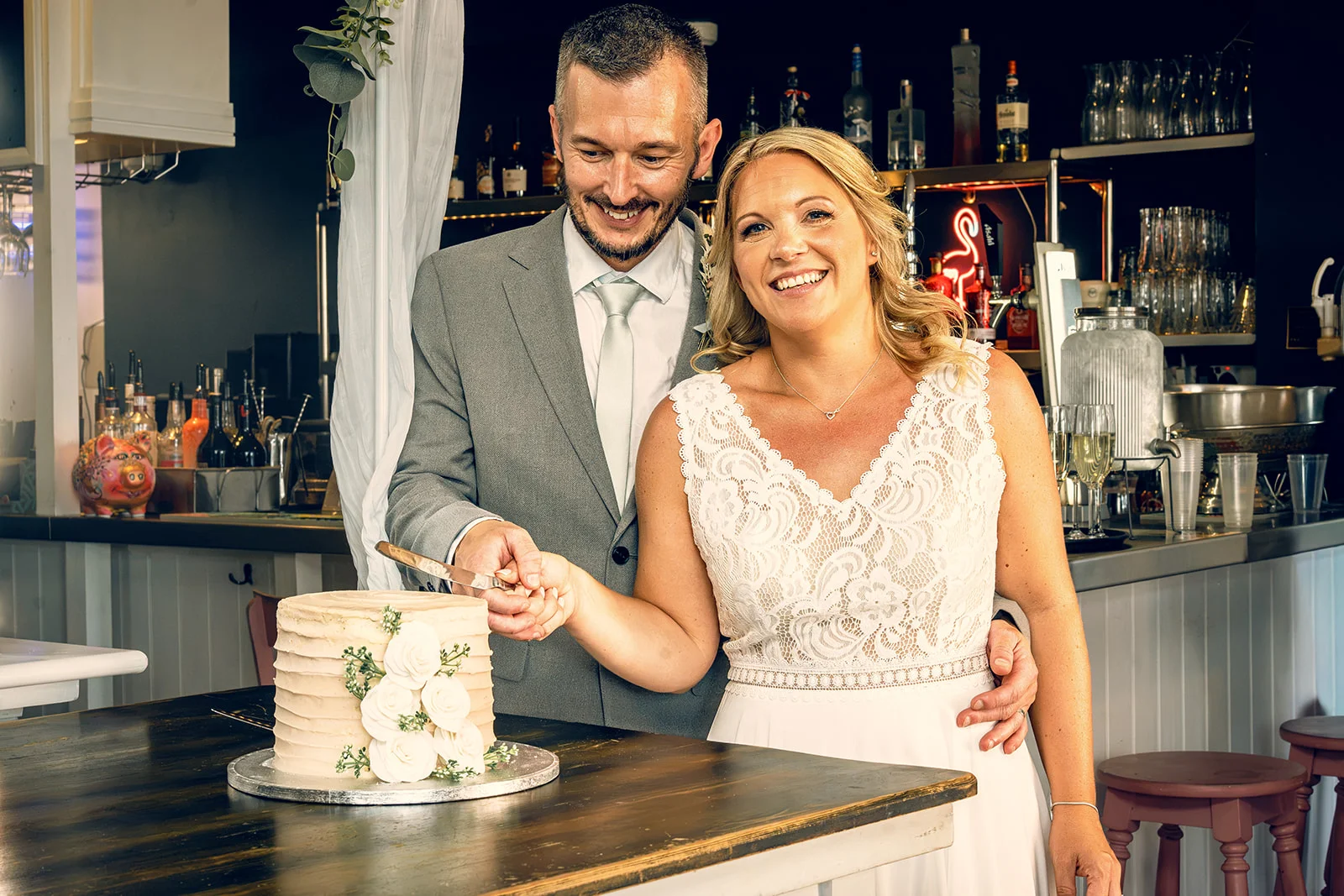 A smiling bride and groom cut a two-tier white textured cake adorned with white roses inside a rustic pub venue.