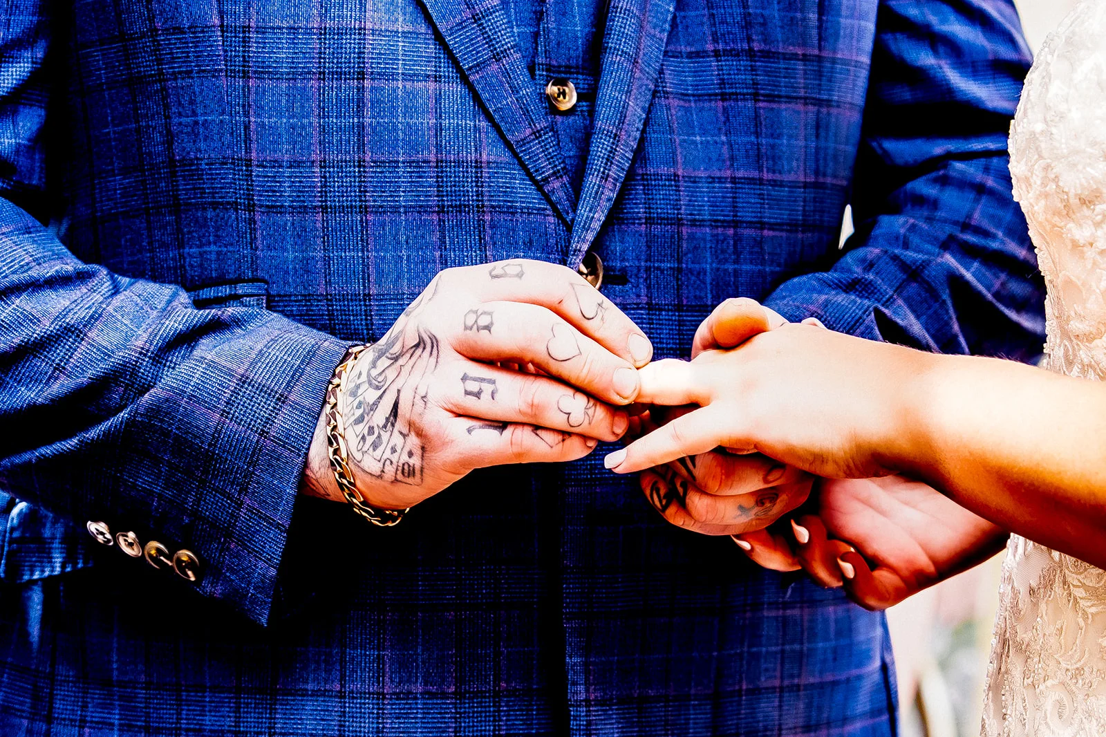 Close-up shot of a groom wearing a blue checked suit sliding a wedding ring onto the bride's finger, showcasing his hand tattoos and a gold chain bracelet.