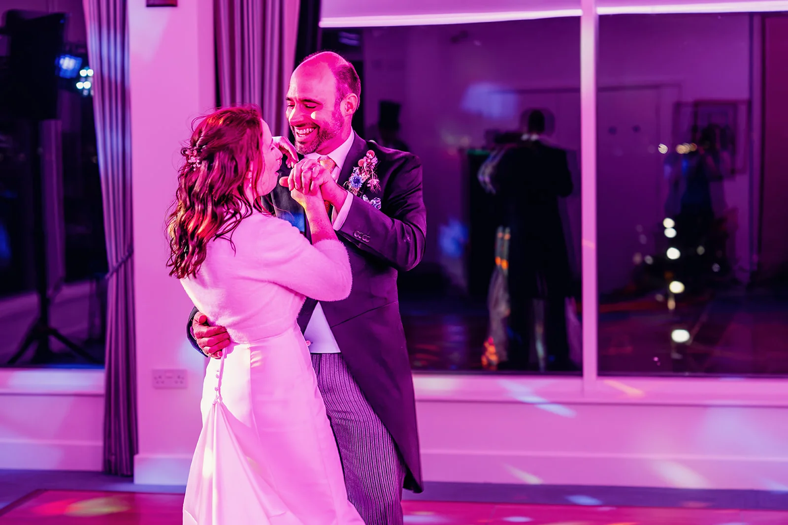 A smiling bride and groom share a joyful first dance together under vibrant pink evening lighting.