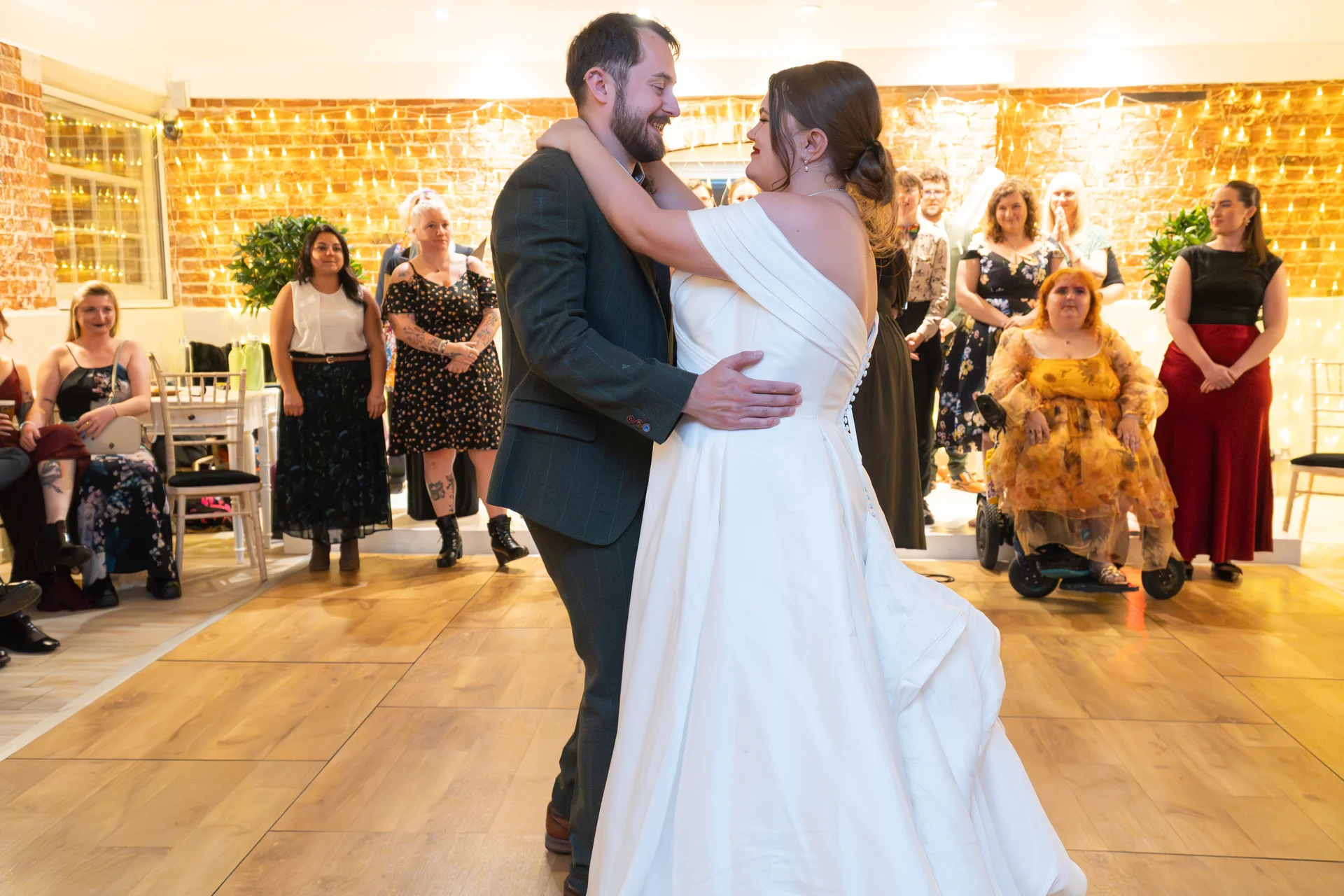 The bride and groom share a joyful first dance on a wooden floor in a venue featuring exposed brick and a warm fairy light canopy.