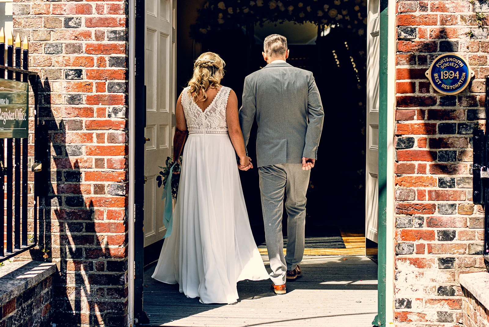 A newlywed couple walks hand in hand away from the camera, entering through open double doors into a red brick registry office building bathed in bright sunlight.