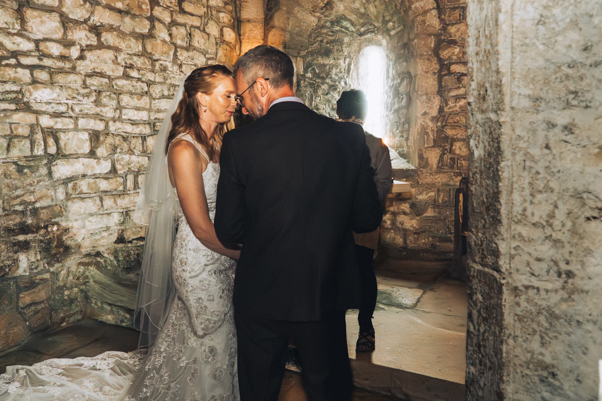 A bride and groom share an intimate nose-to-nose moment inside a historic stone church with exposed walls and warm window light.