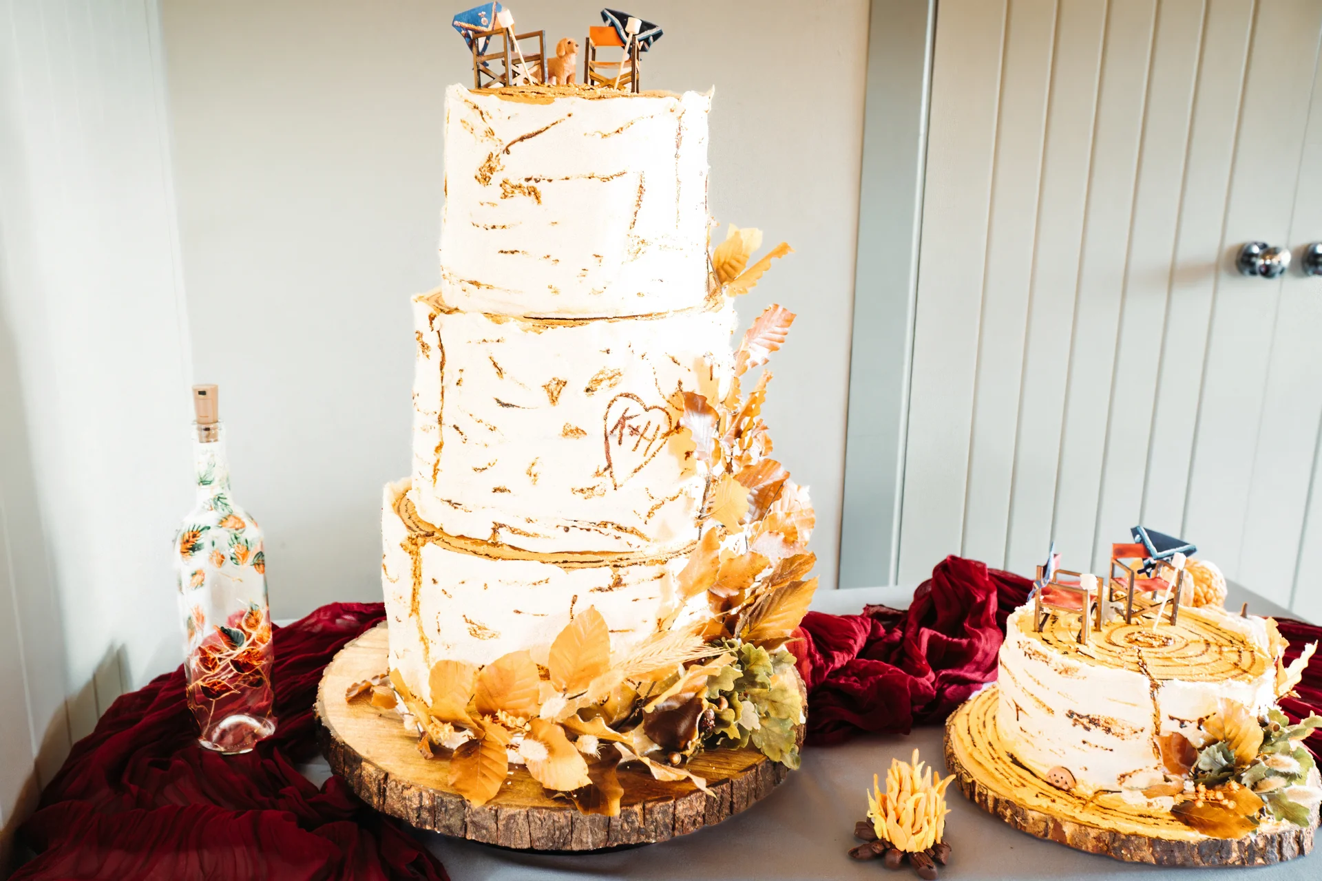 A stunning three-tiered rustic wedding cake styled to resemble birch bark features golden autumn leaf cascading details, displayed on a wooden slice stand next to a smaller single-tier cake and a bottle of wine on a burgundy cloth.