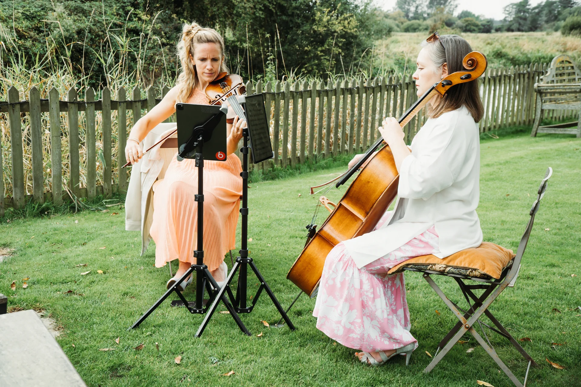 A violinist and a cellist perform live music outdoors in a green garden setting during a wedding ceremony.