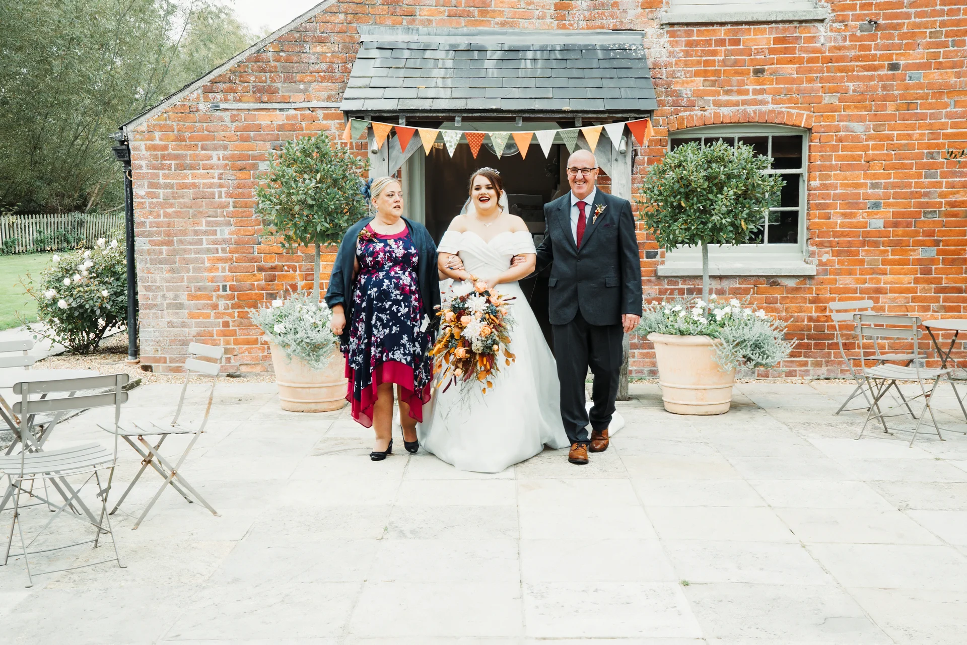 A laughing bride flanked by her mother and father walks out from a rustic red brick barn adorned with colourful bunting during a joyful wedding celebration.