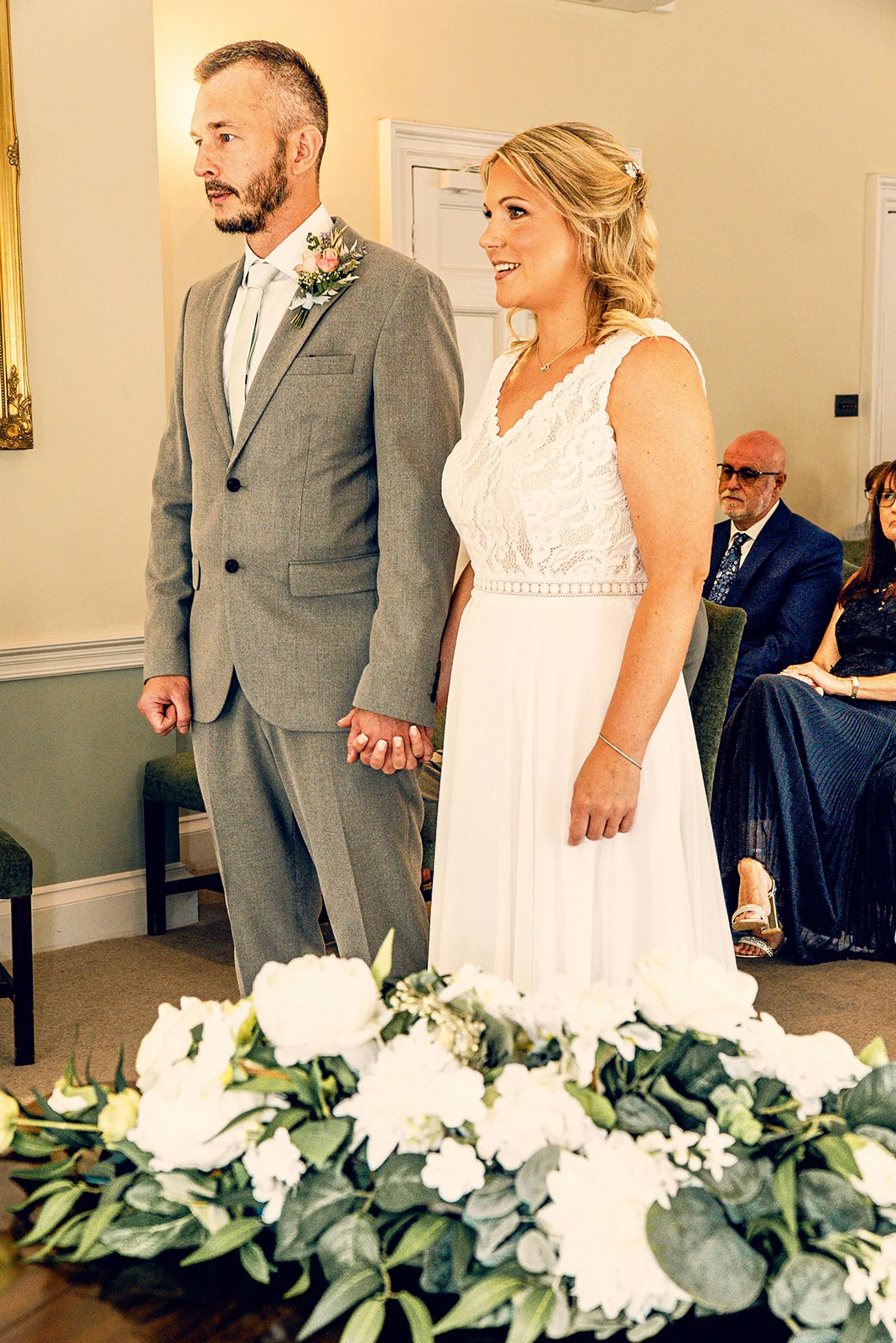 A smiling bride in a lace-bodice wedding gown and a groom in a grey suit hold hands during an indoor civil ceremony, framed by white floral arrangements in the foreground.