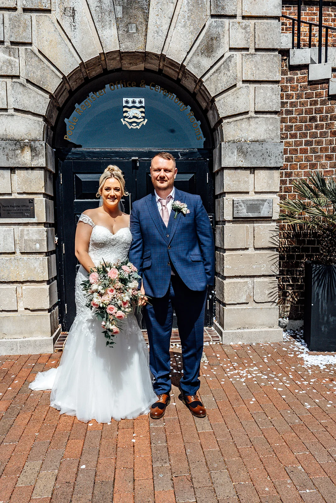 A newly married couple poses outside a registry office entrance with stone archways, featuring the bride in a lace gown holding a cascading rose bouquet and the groom in a blue checked suit.