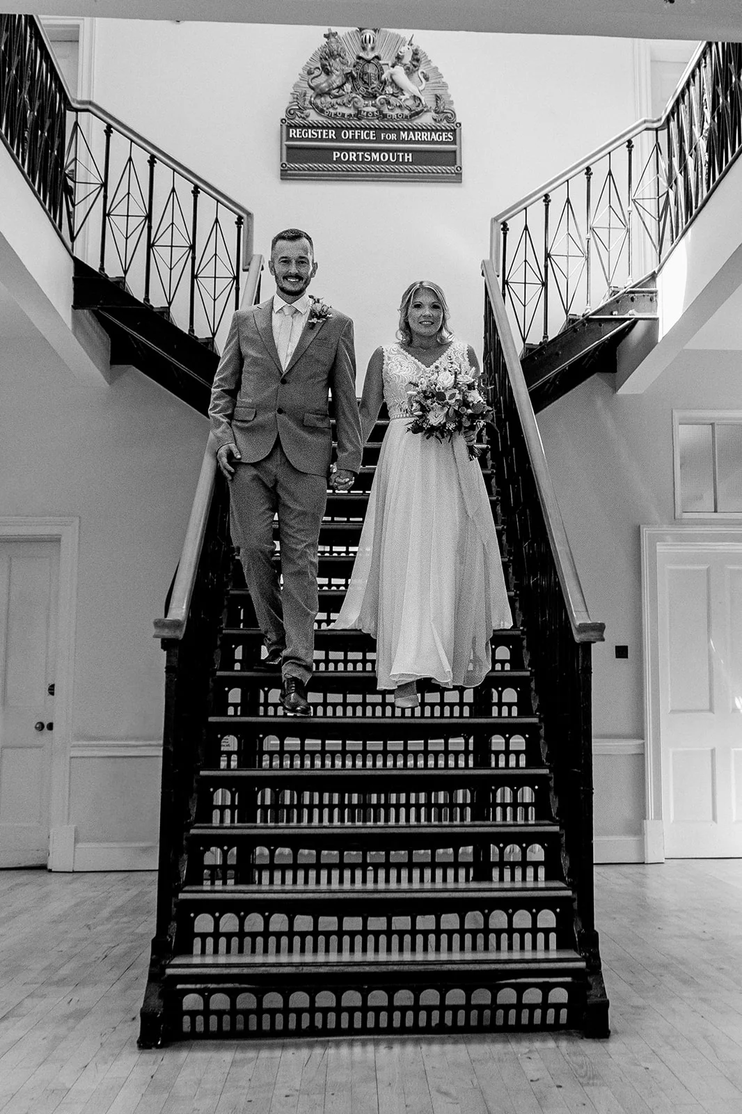 A newlywed couple happily walks hand in hand down a grand staircase inside a registry office, with the bride holding a bouquet.