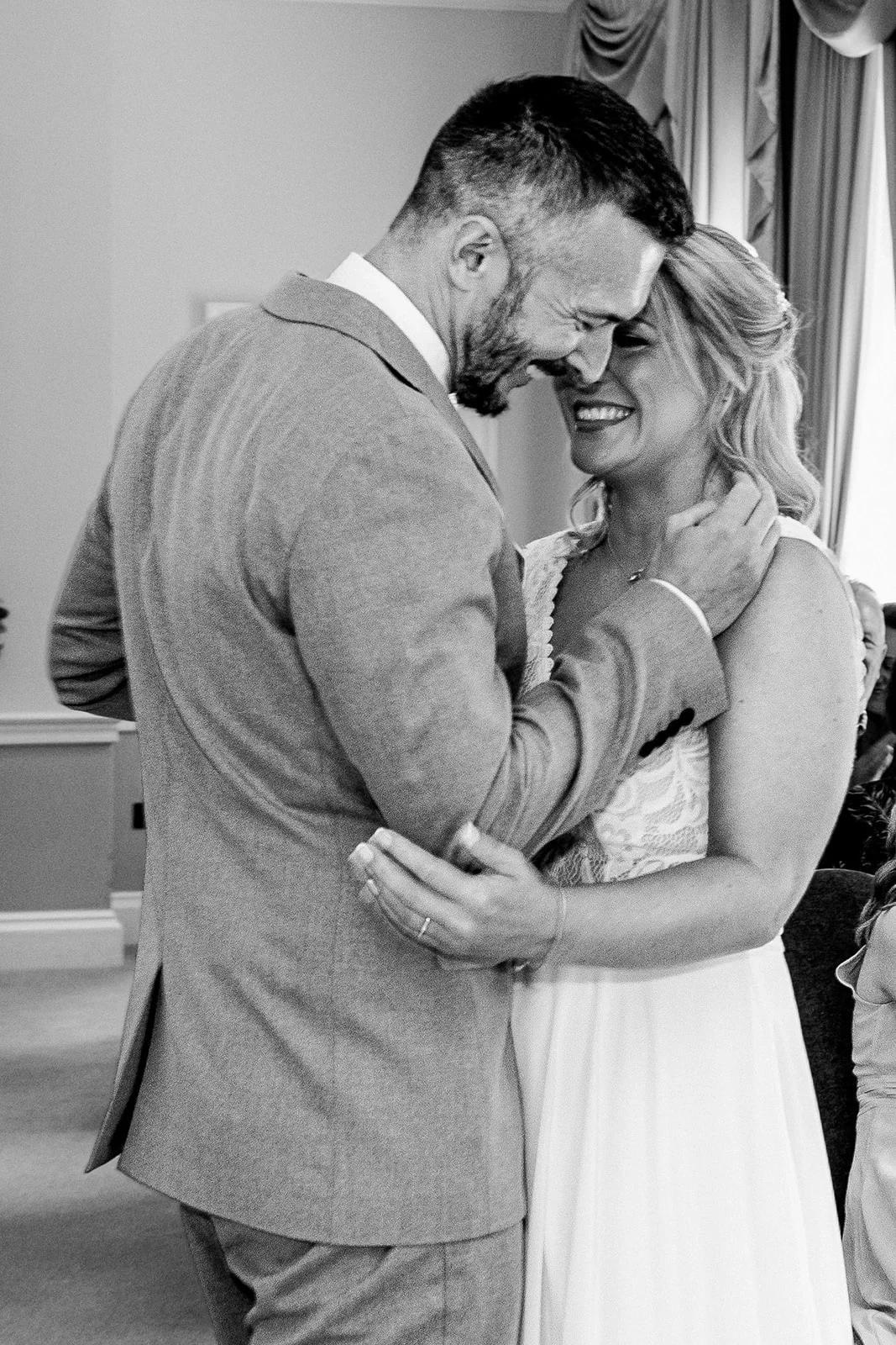 A joyous bride and groom embrace nose to nose, laughing happily together during their wedding celebration captured in a candid black and white shot.