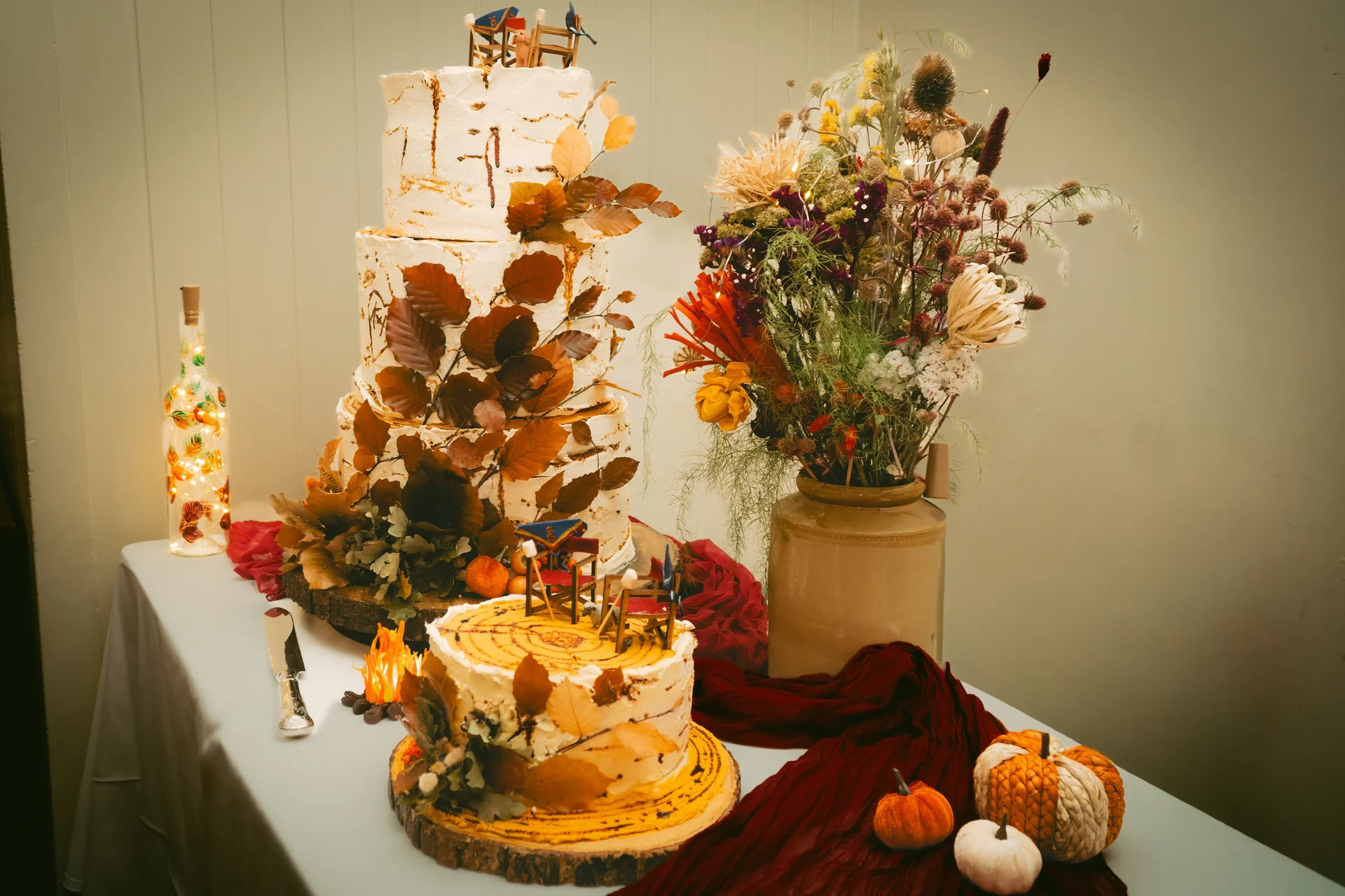 A rustic wedding cake table featuring a tiered cake and a smaller cake adorned with autumn leaves, accompanied by a dried flower arrangement and miniature pumpkins.