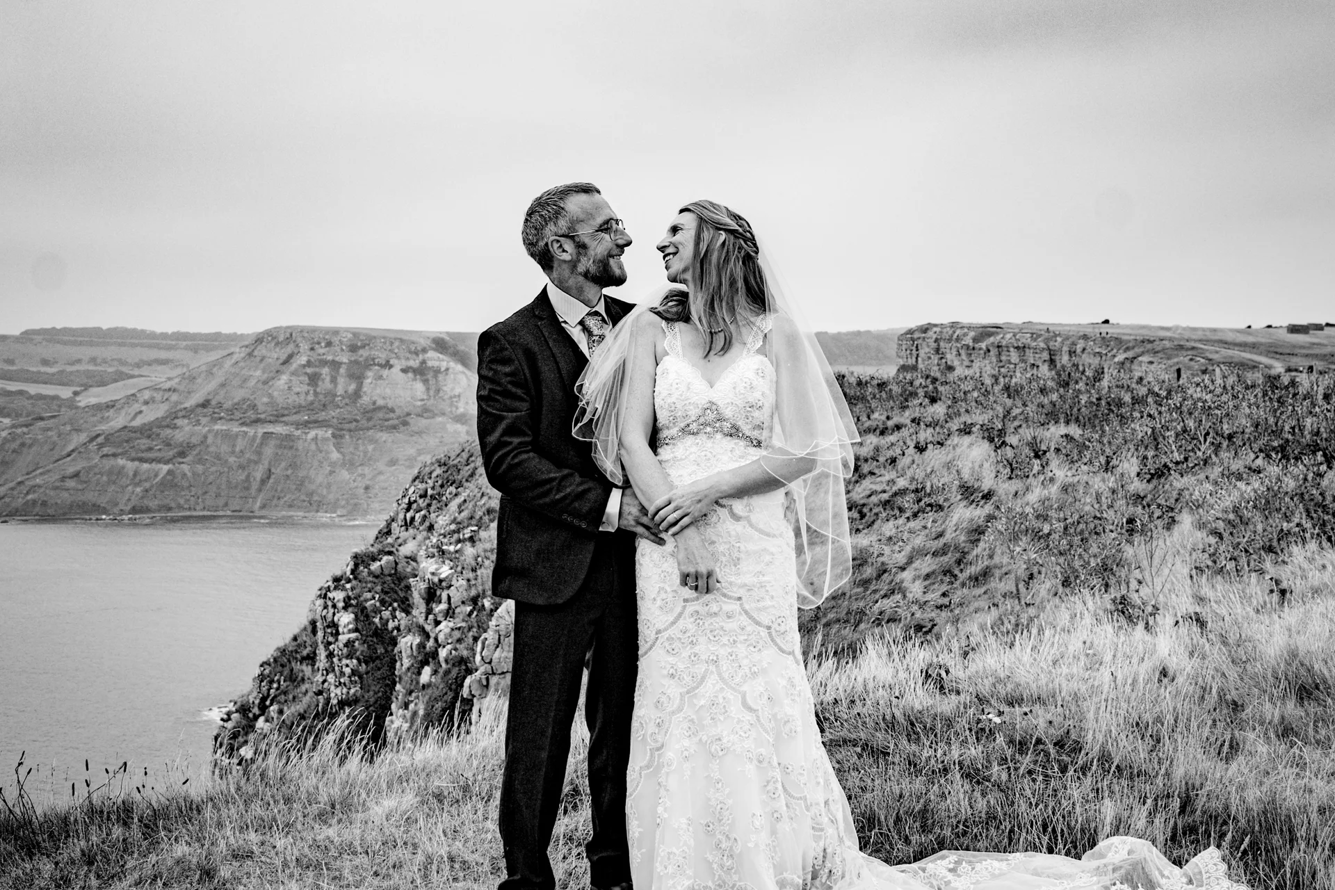 A newlywed couple shares a loving glance while standing on a dramatic clifftop overlooking the sea during their wedding portrait session.