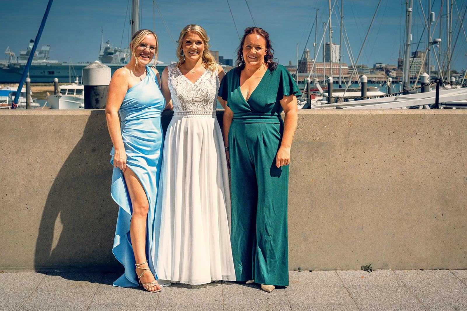 A bride in a white lace and pleated gown stands outdoors by a marina alongside two wedding party members in blue and green attire.