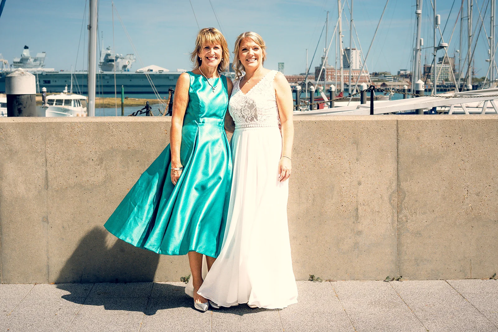 A mother of the bride in a vibrant teal dress and the bride in a lace and chiffon gown pose happily together along a harbour promenade on a bright sunny day.