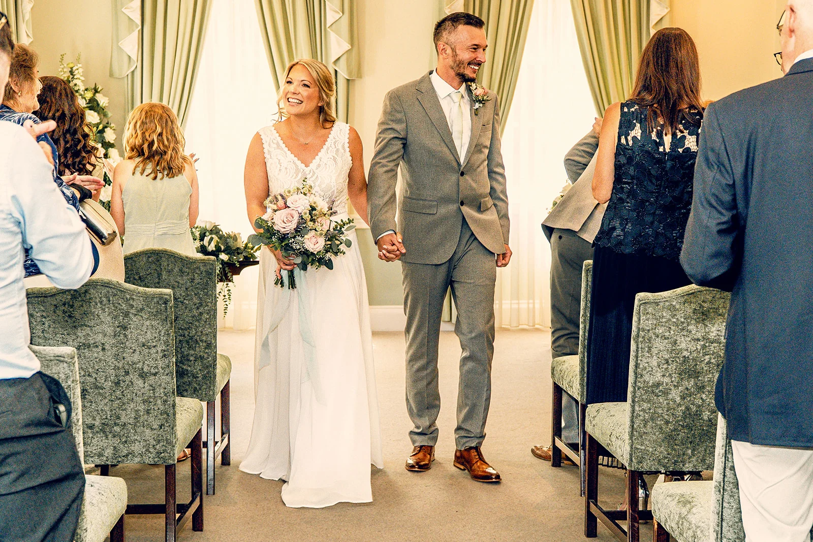 A smiling bride and groom walk hand in hand down the aisle after their ceremony, cheered on by seated wedding guests.