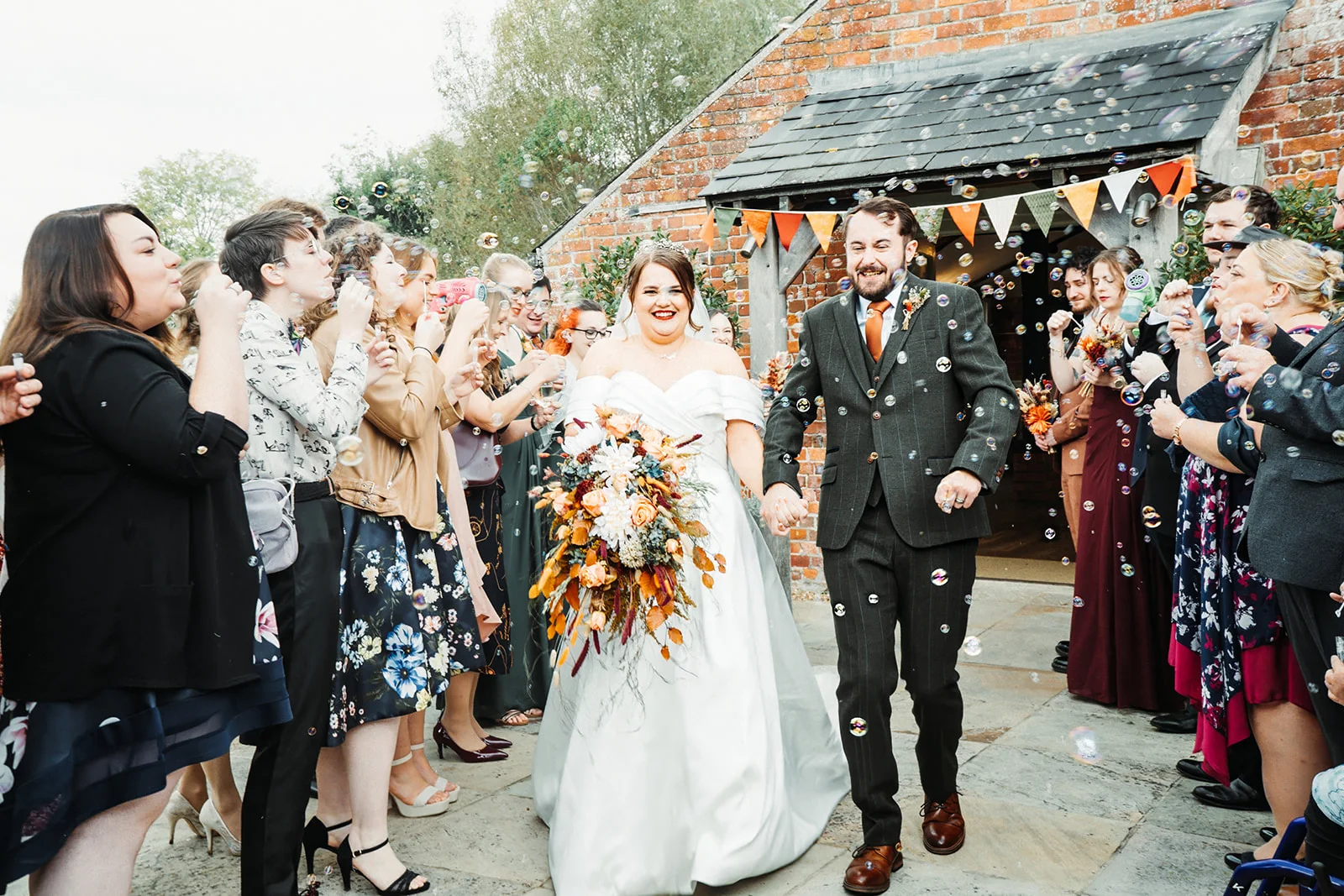 A joyful bride and groom walk hand in hand through a bubble exit held by cheering guests outside a rustic brick barn adorned with colourful bunting.