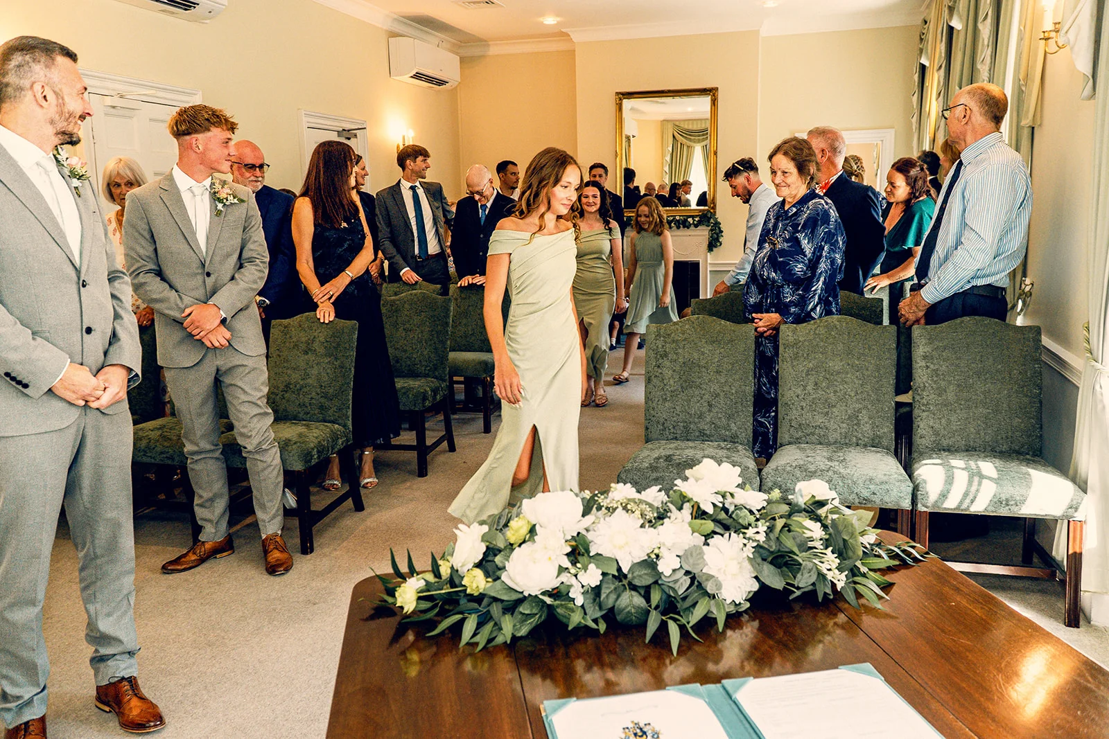 A bridesmaid in a sage green off-the-shoulder dress walks down the aisle towards the ceremony table as guests watch from seated chairs.