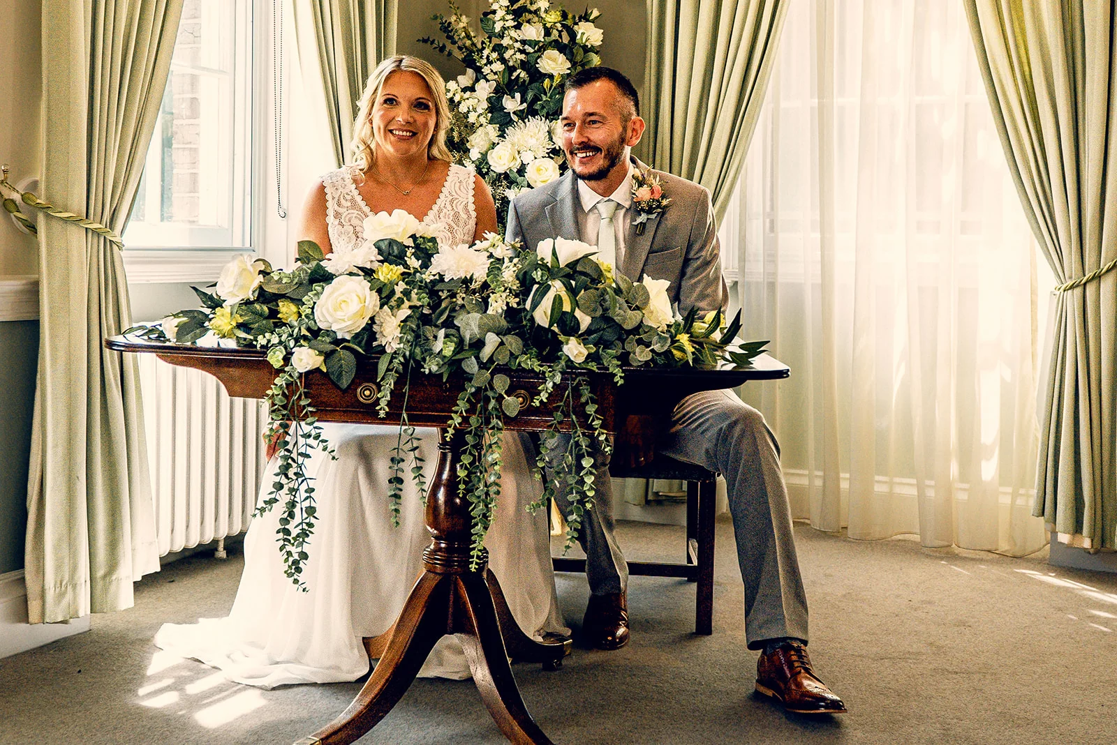 A smiling bride and groom sit together at a wooden table adorned with white and green floral arrangements while signing the register during their civil ceremony.