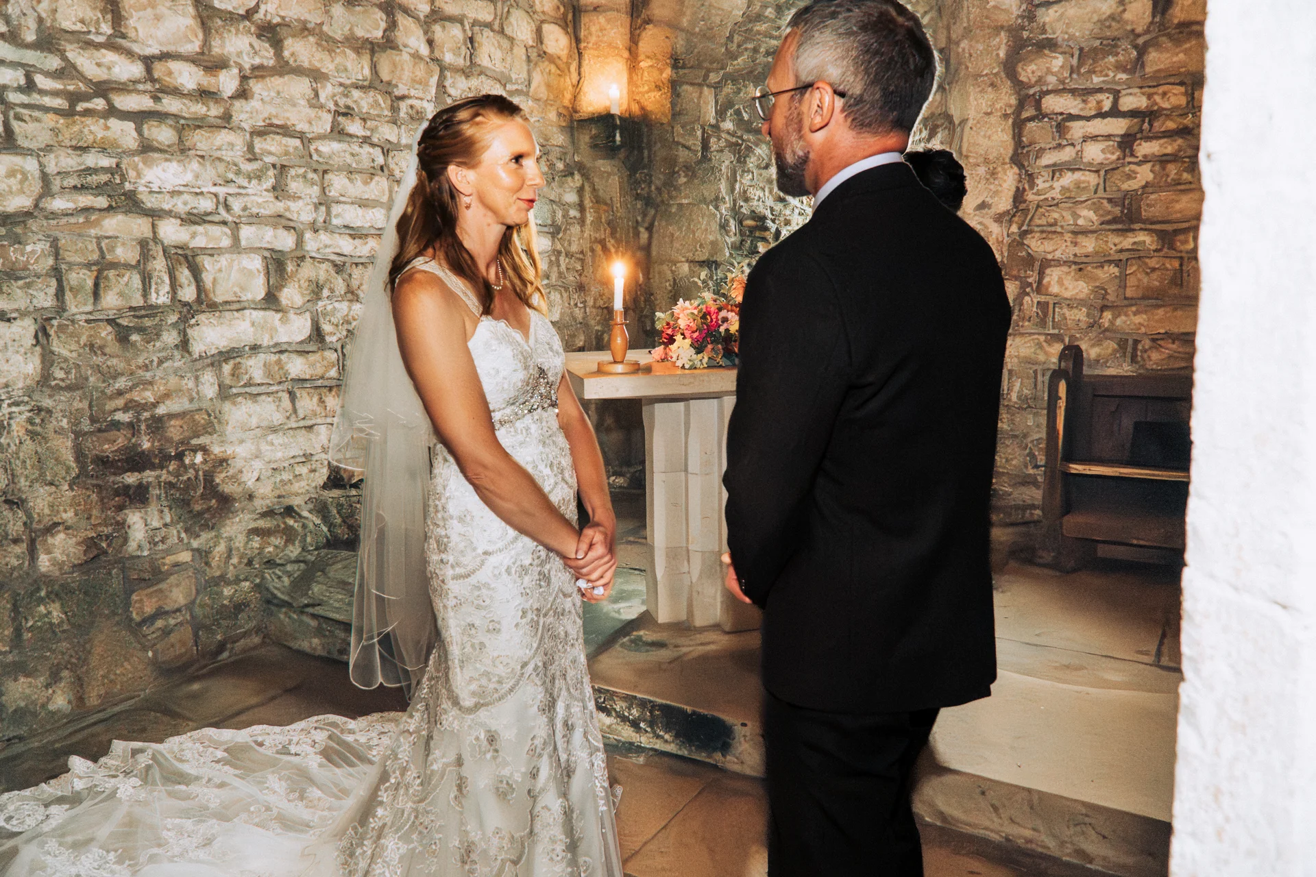 A bride in a detailed lace gown and veil stands facing her groom during an intimate ceremony inside an atmospheric stone chapel illuminated by candlelight.