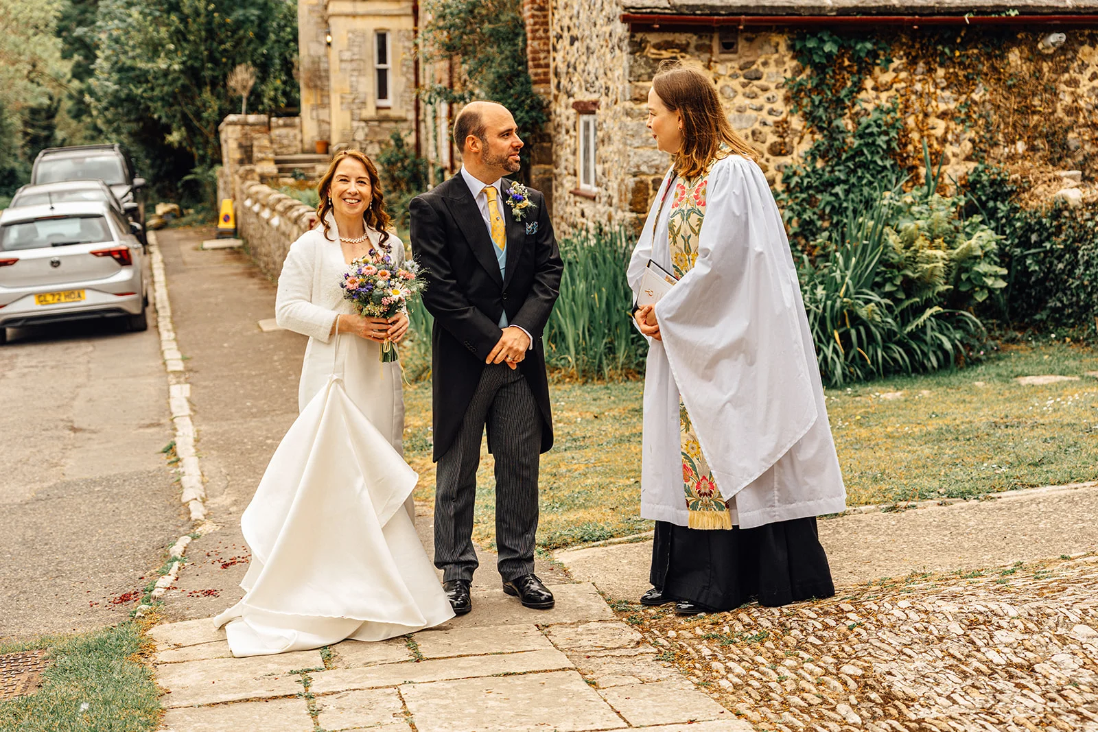 A bride holding a wildflower bouquet and a groom in a morning suit stand outside a stone church building talking to an officiant in religious vestments.