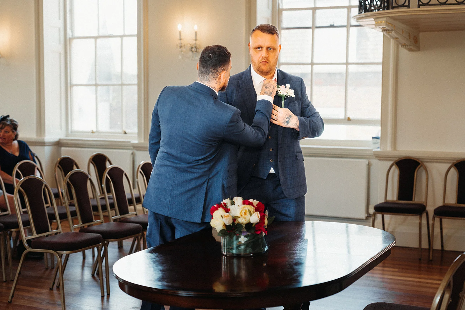 A groom in a navy suit adjusts his tie with the help of his partner inside a bright registry office with rows of chairs.