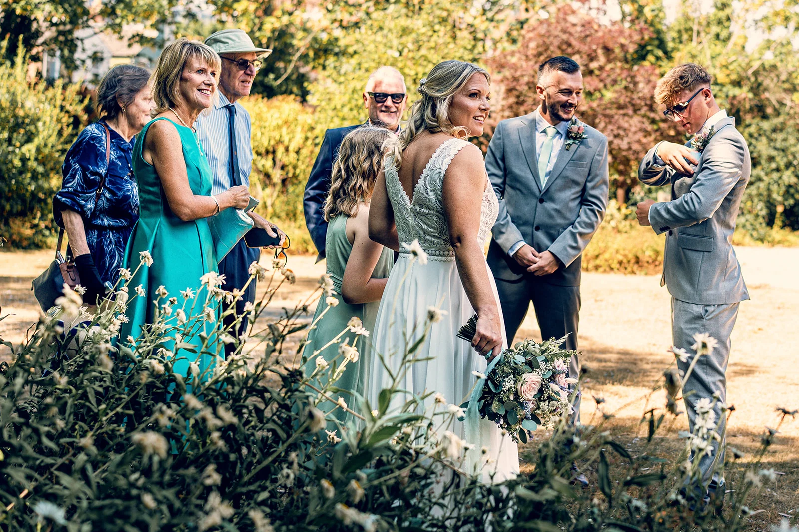 A bride holding a hand-tied floral bouquet stands smiling outdoors with her groom and wedding guests in a sunlit garden setting.