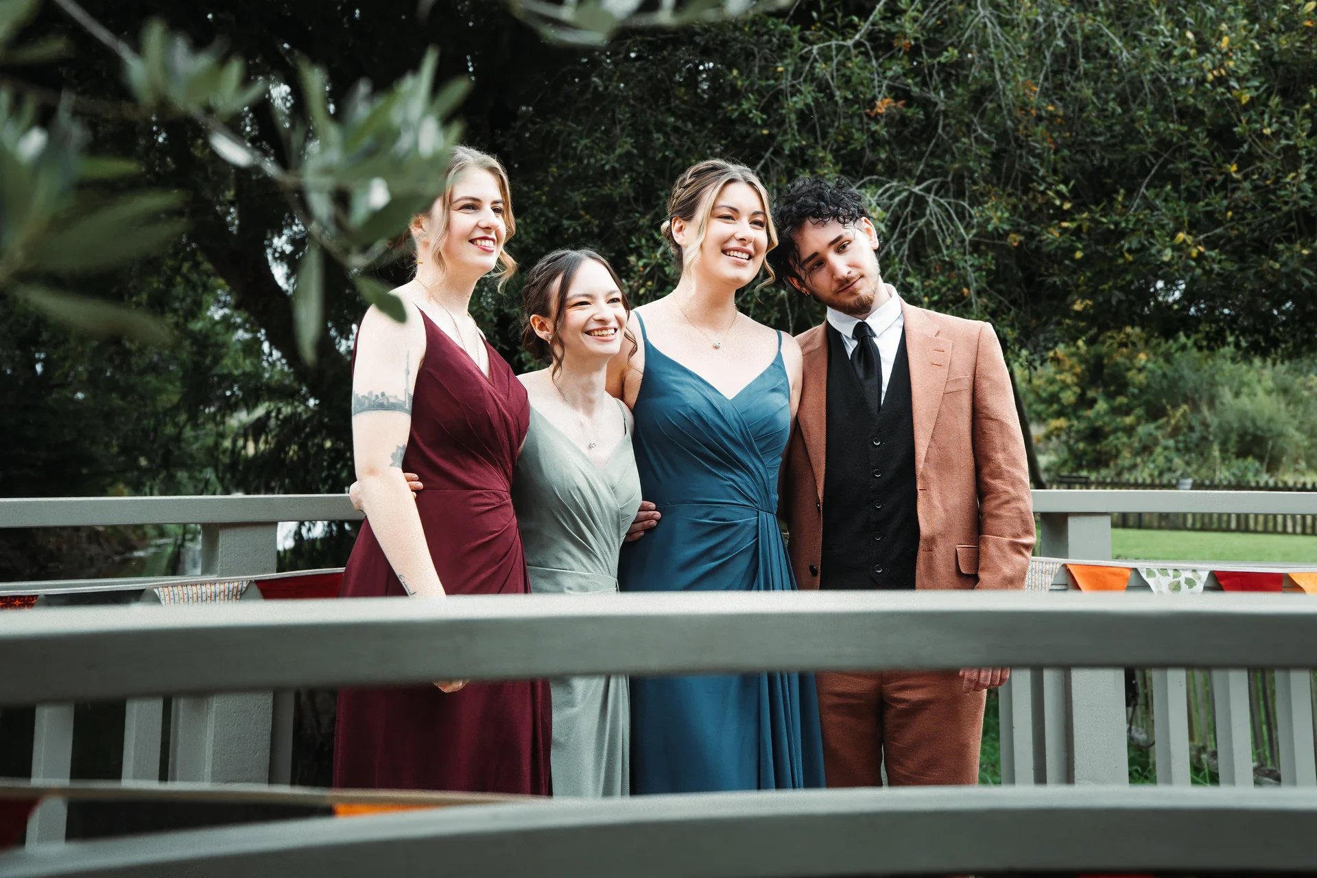 A wedding party of three bridesmaids in mismatched jewel-toned dresses and a groomsman in a tan suit stand smiling together outdoors behind a wooden railing.
