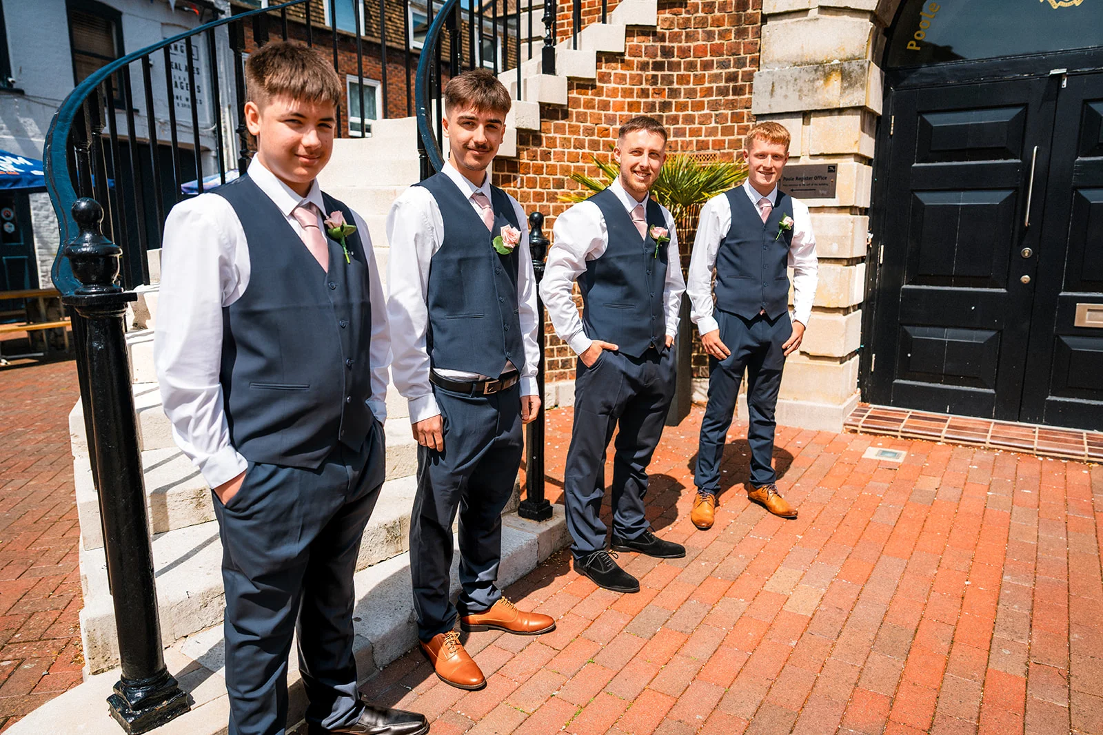 Four groomsmen in matching navy suits, waistcoats and pink ties pose outside a registry office entrance on a sunny day.