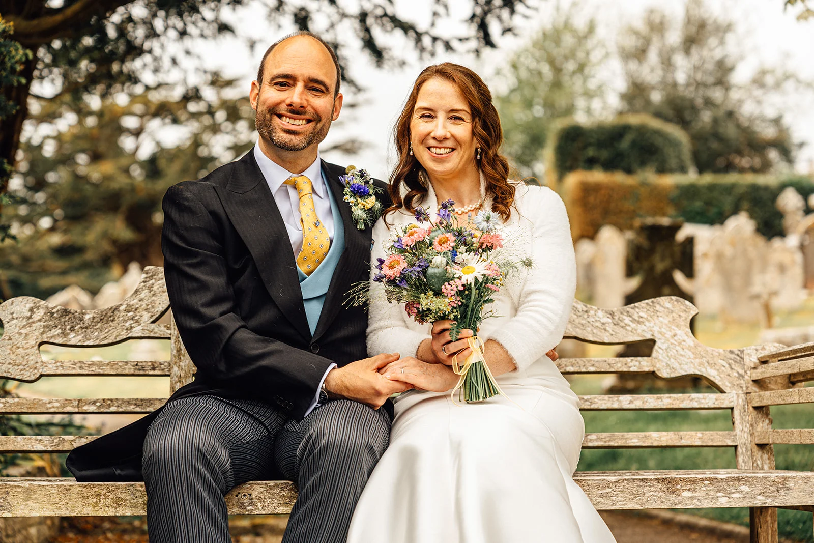 A smiling bride and groom sit together on a weathered wooden garden bench, with the bride holding a colourful wildflower bouquet.