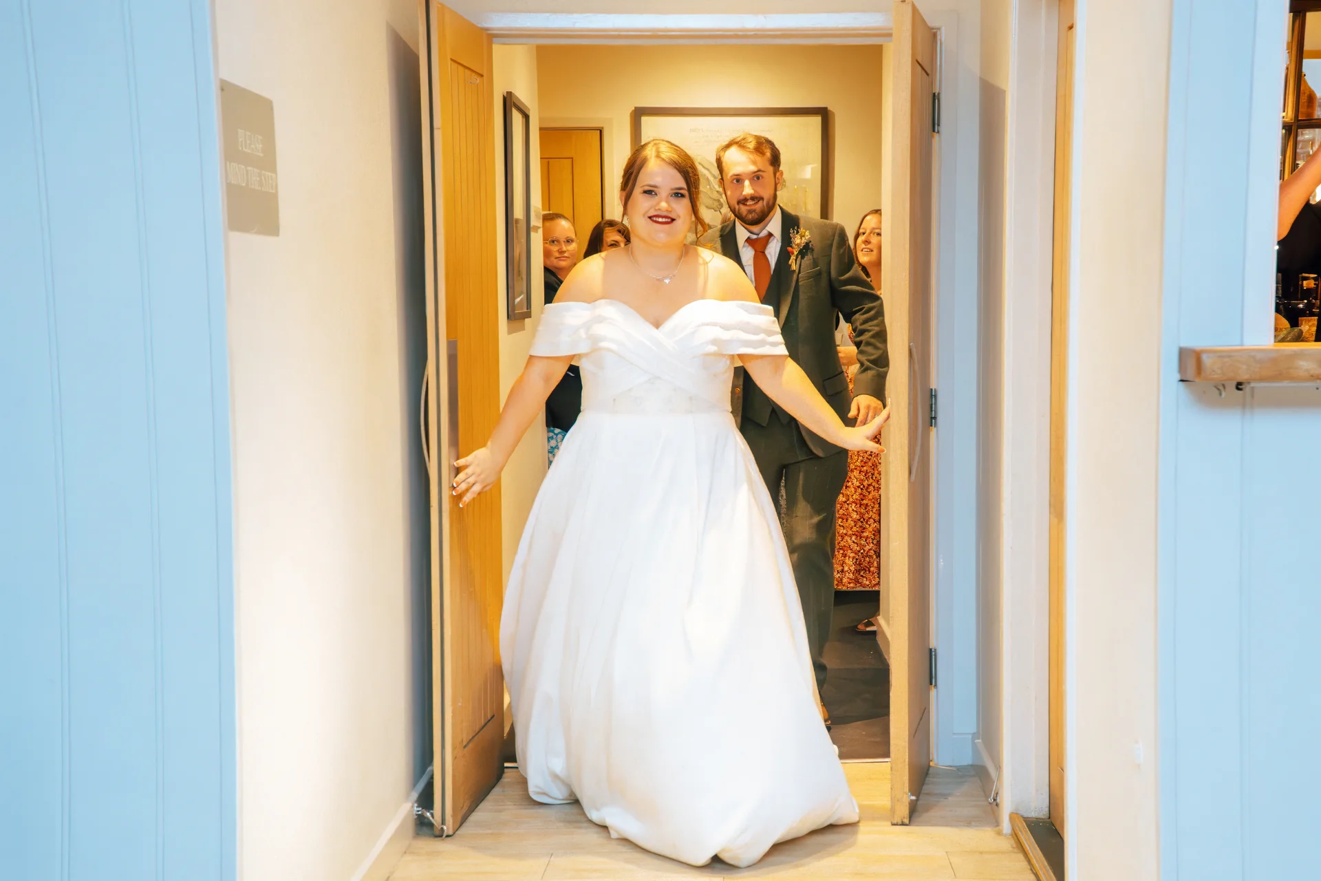 The beaming newlywed couple open a set of wooden doors to enter their wedding reception, with the bride wearing an elegant off-the-shoulder satin gown and the groom in a smart green suit.